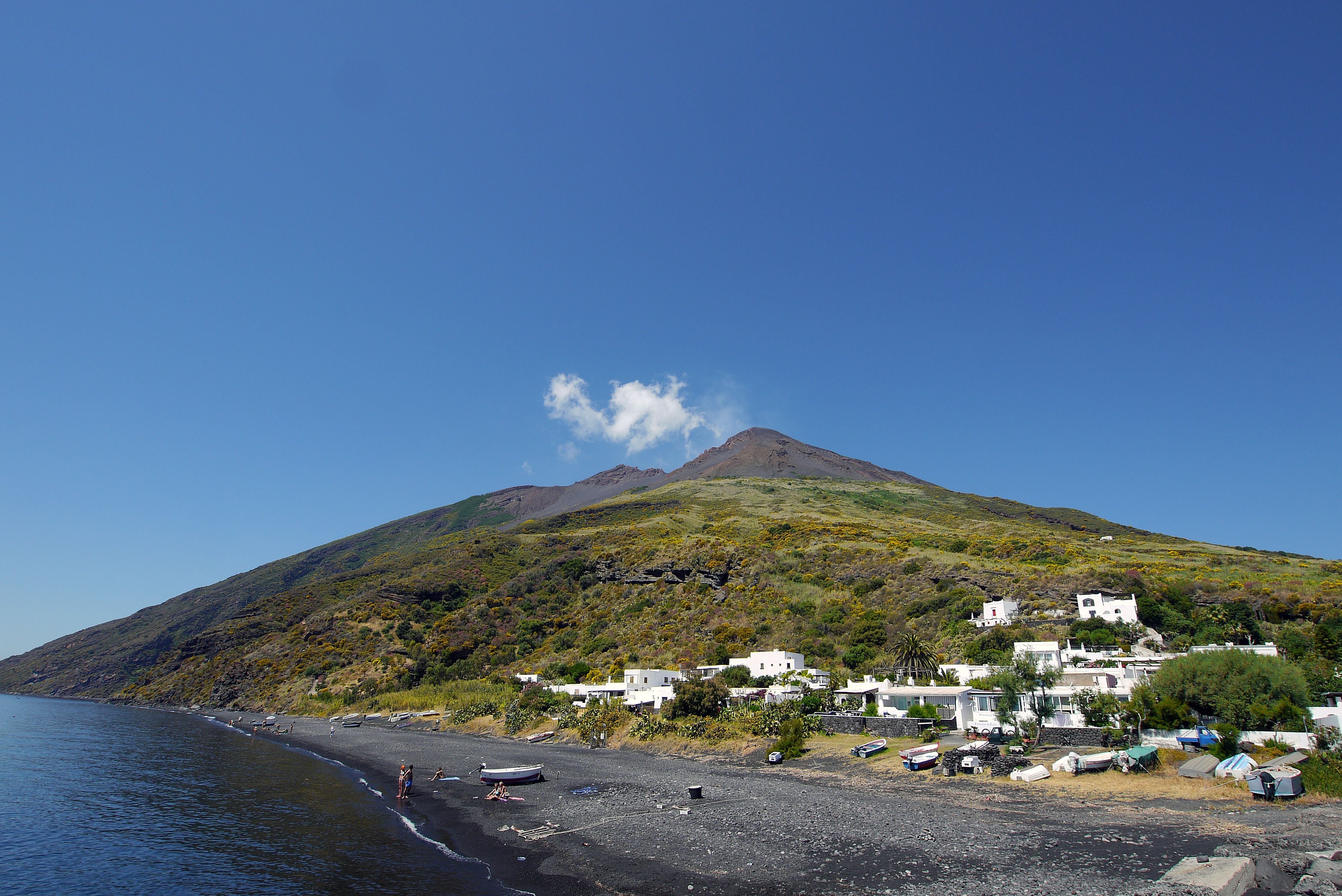 Stromboli Vulkan mit schwarzem Strand, weißen Häusern und Fischerbooten bei Sonnenschein.