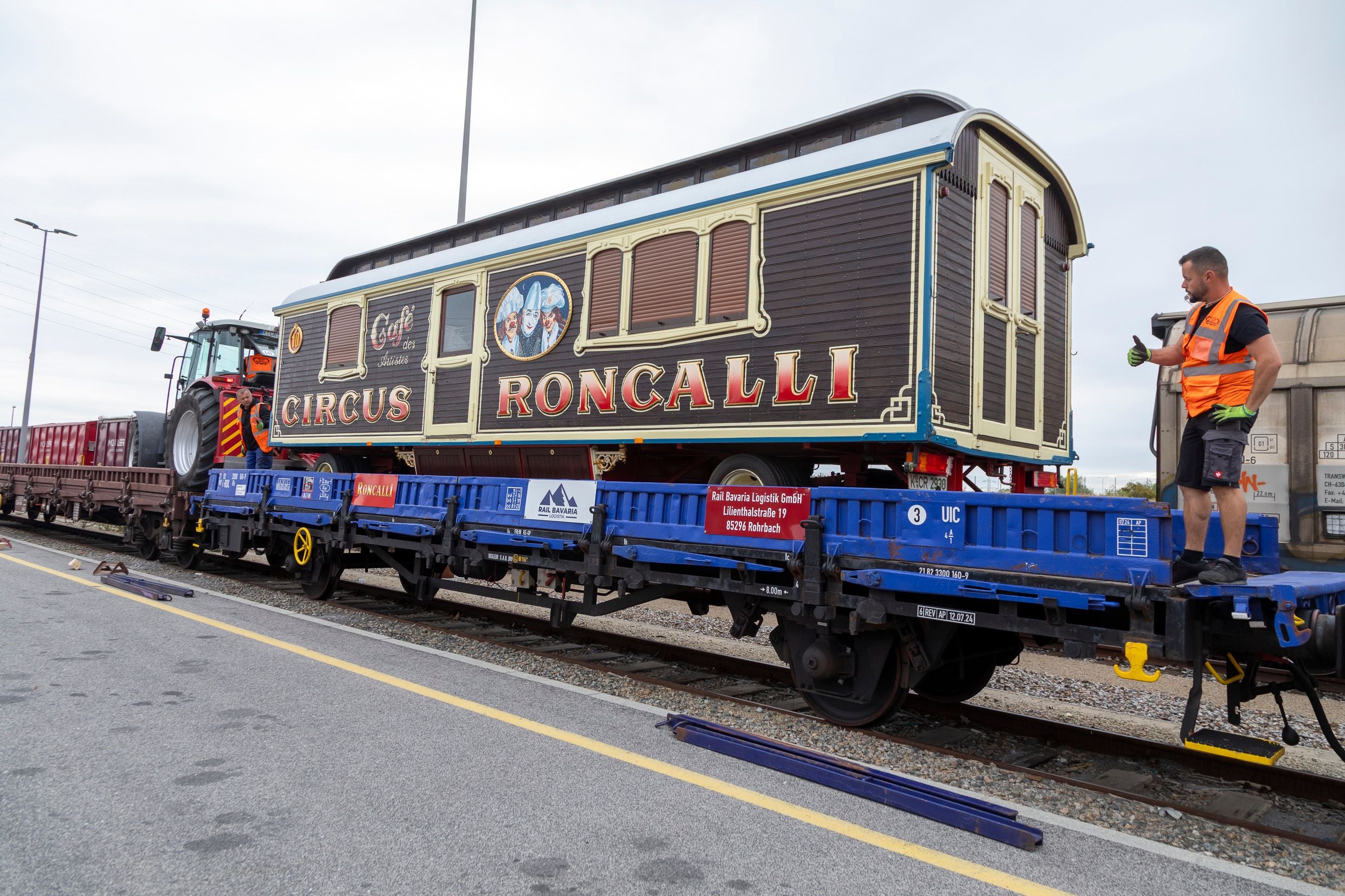 Ein Zirkuswagen von Circus Roncalli steht auf einem blauen Eisenbahnwaggon, daneben ein Arbeiter.