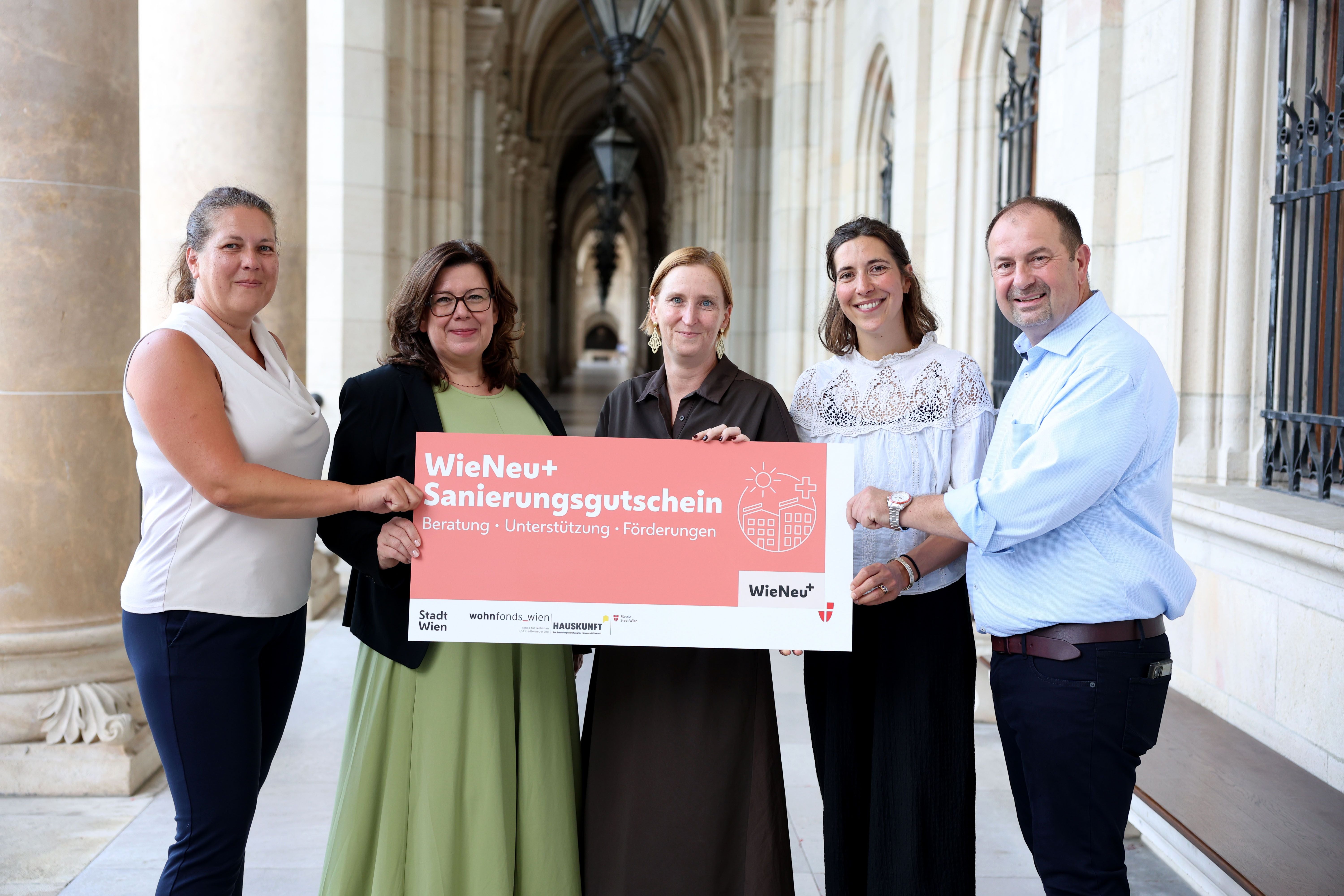 Fünf Personen halten gemeinsam ein Schild mit der Aufschrift „Sanierungsgutschein“ in einem Säulengang.