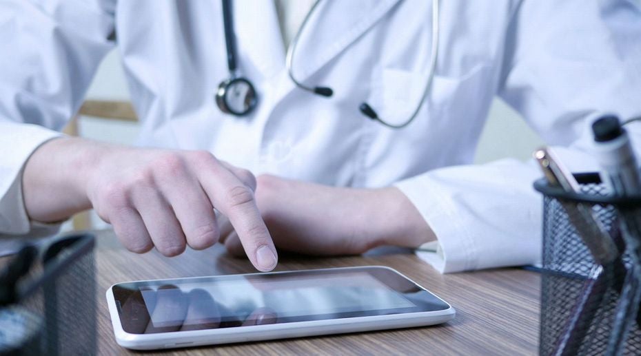 Arzt im weißen Kittel bedient ein Tablet auf einem Schreibtisch mit Stethoskop um den Hals.