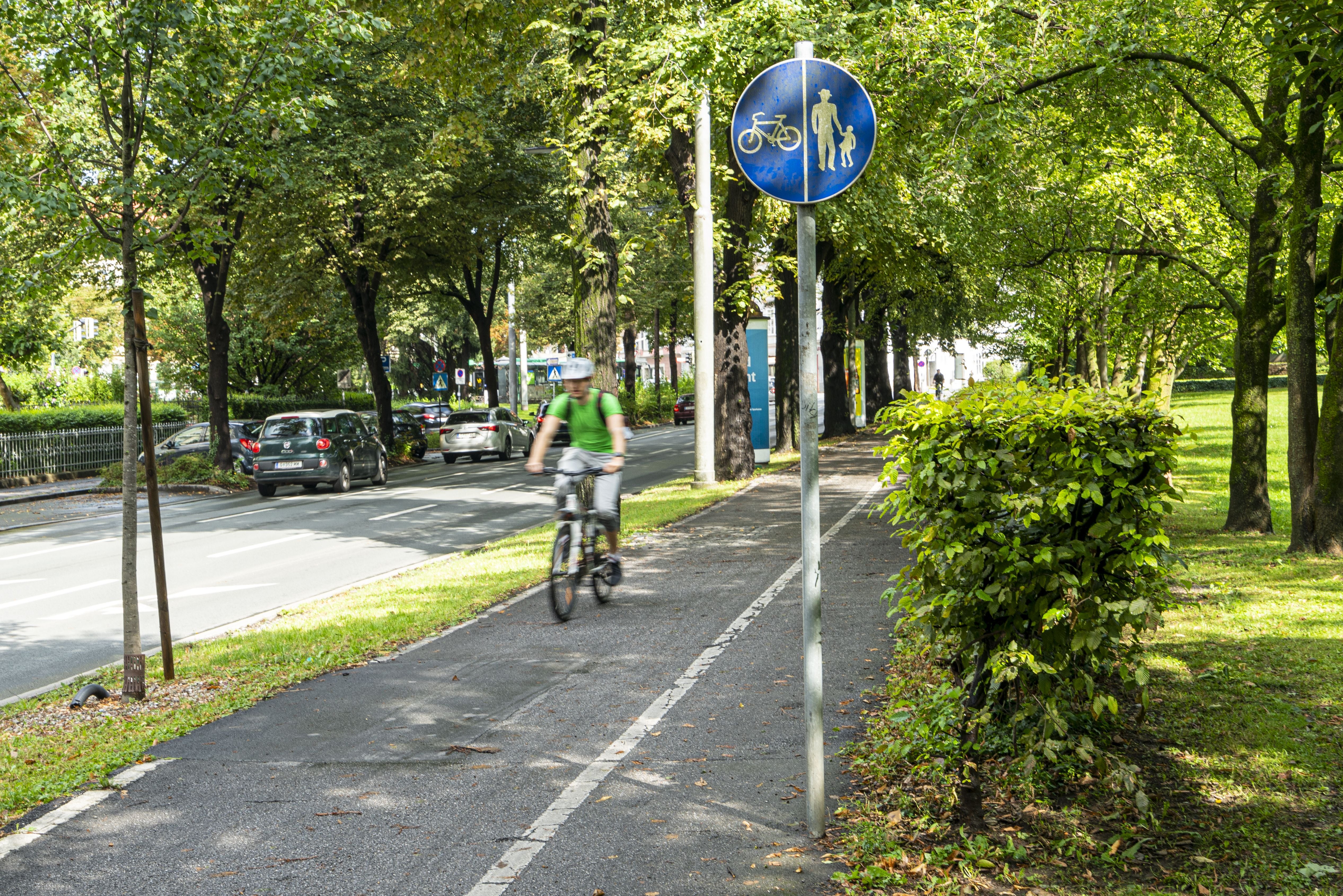 Radweg im Stadtpark Graz mit Verkehrsschild, Radfahrer und grünen Bäumen im Sommer.