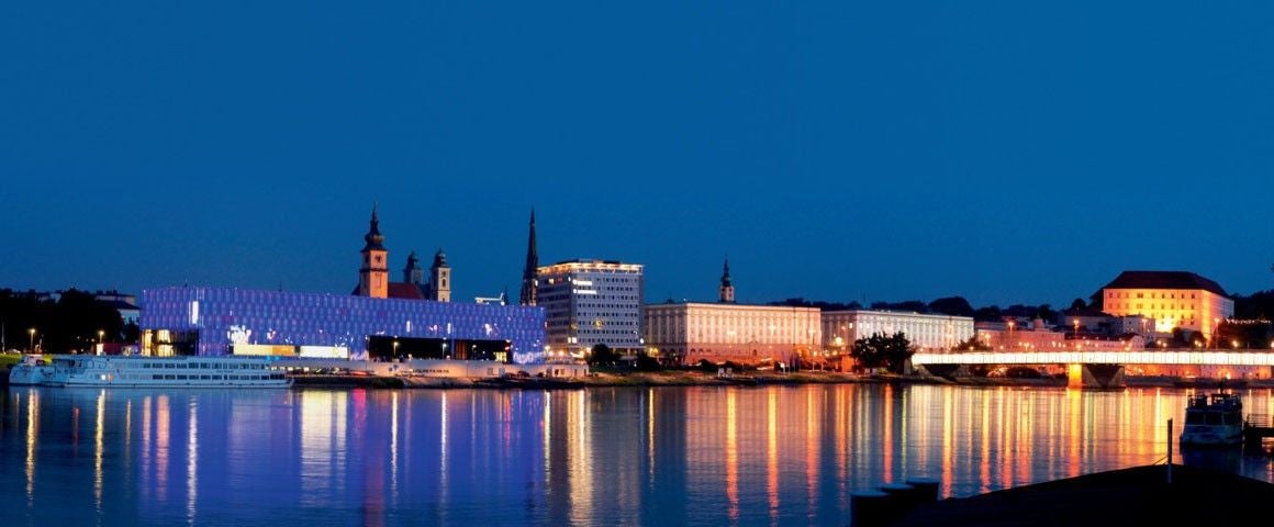Die beleuchtete Skyline von Linz spiegelt sich abends im ruhigen Wasser der Donau.