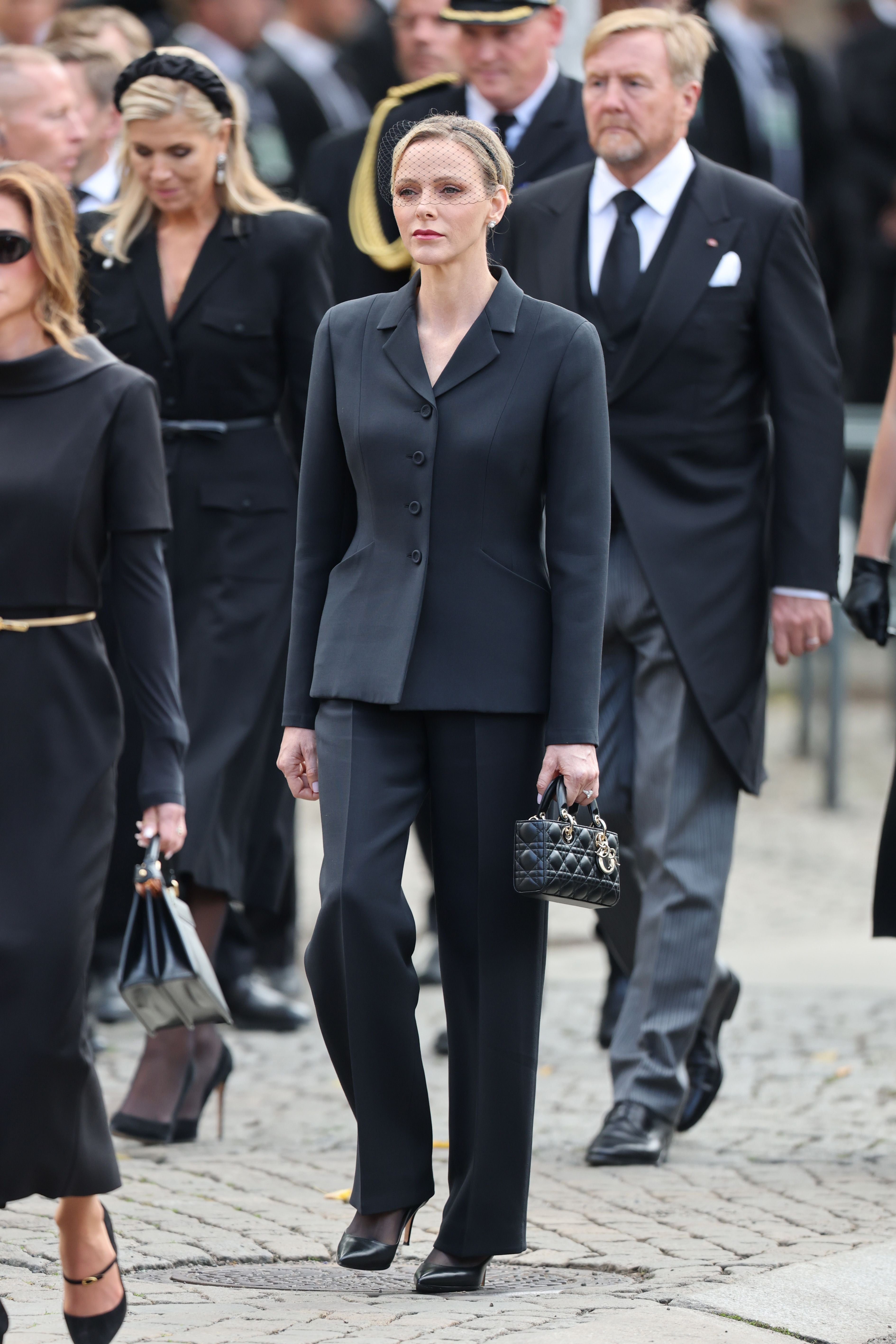 Eine Frau in schwarzem Anzug und Schleier hält eine kleine schwarze Handtasche bei einer Trauerfeier.