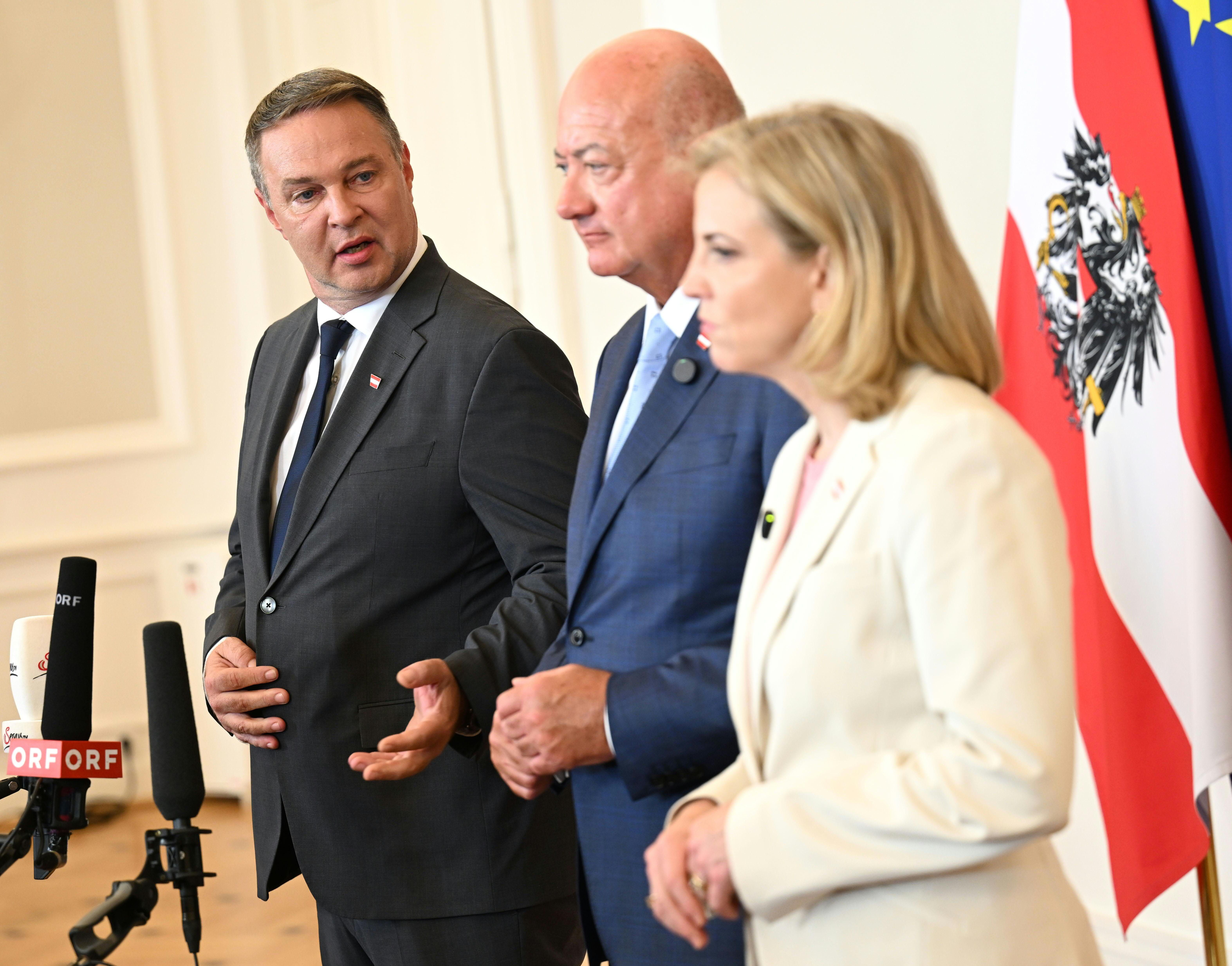 Drei Politiker bei einer Pressekonferenz mit Mikrofonen und österreichischer Flagge im Hintergrund.