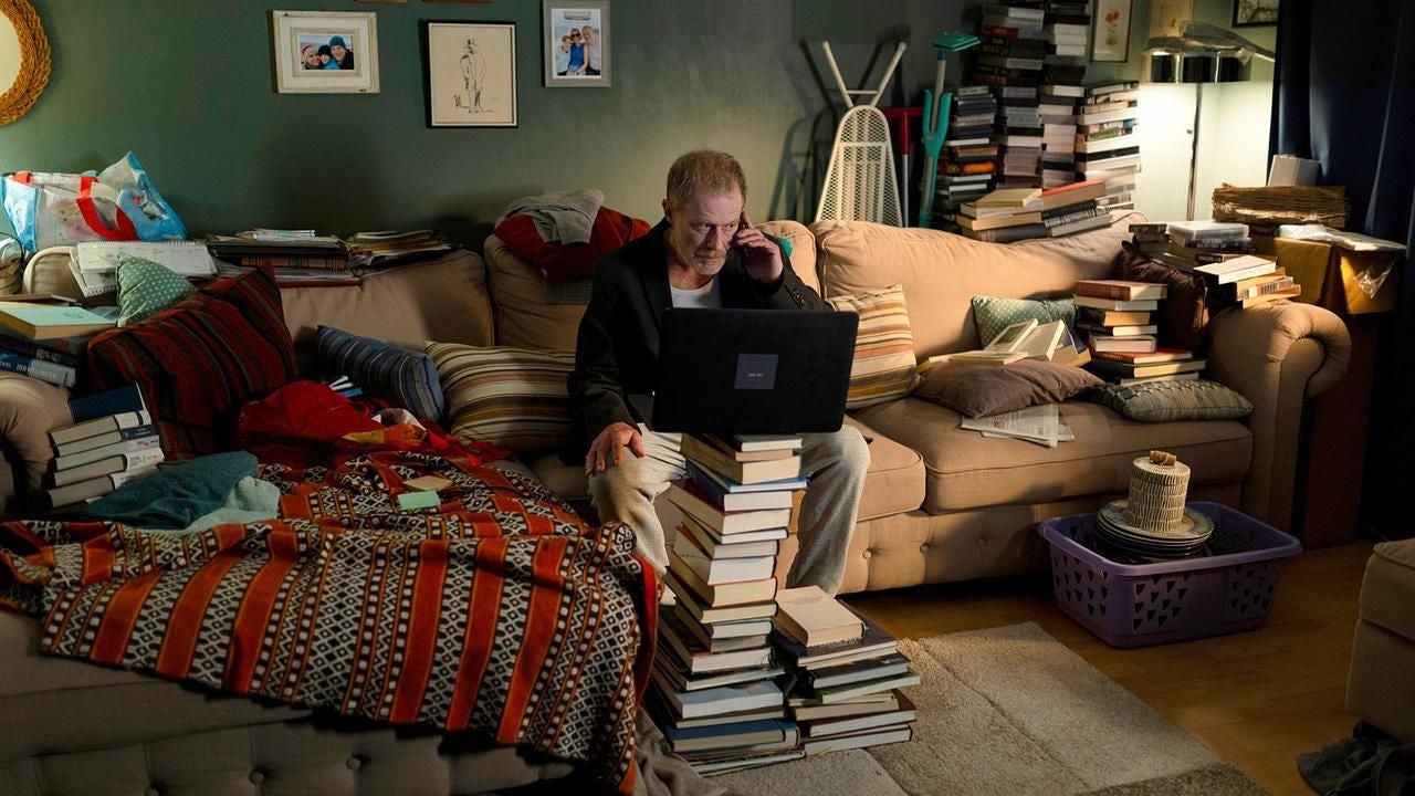 Ein Mann sitzt in einem unordentlichen Wohnzimmer vor einem Laptop, umgeben von vielen Büchern.