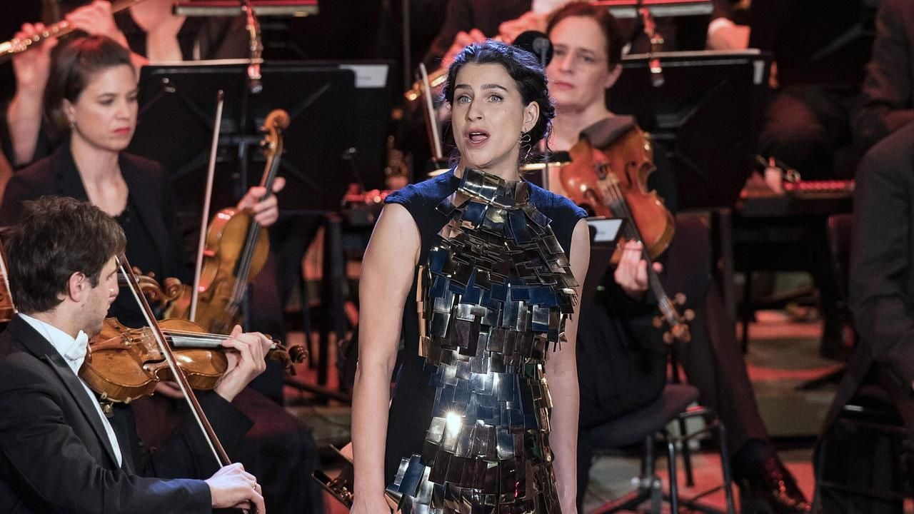 Eine Sängerin mit extravagantem Kleid steht vor einem Orchester aus Streich- und Blasinstrumenten.
