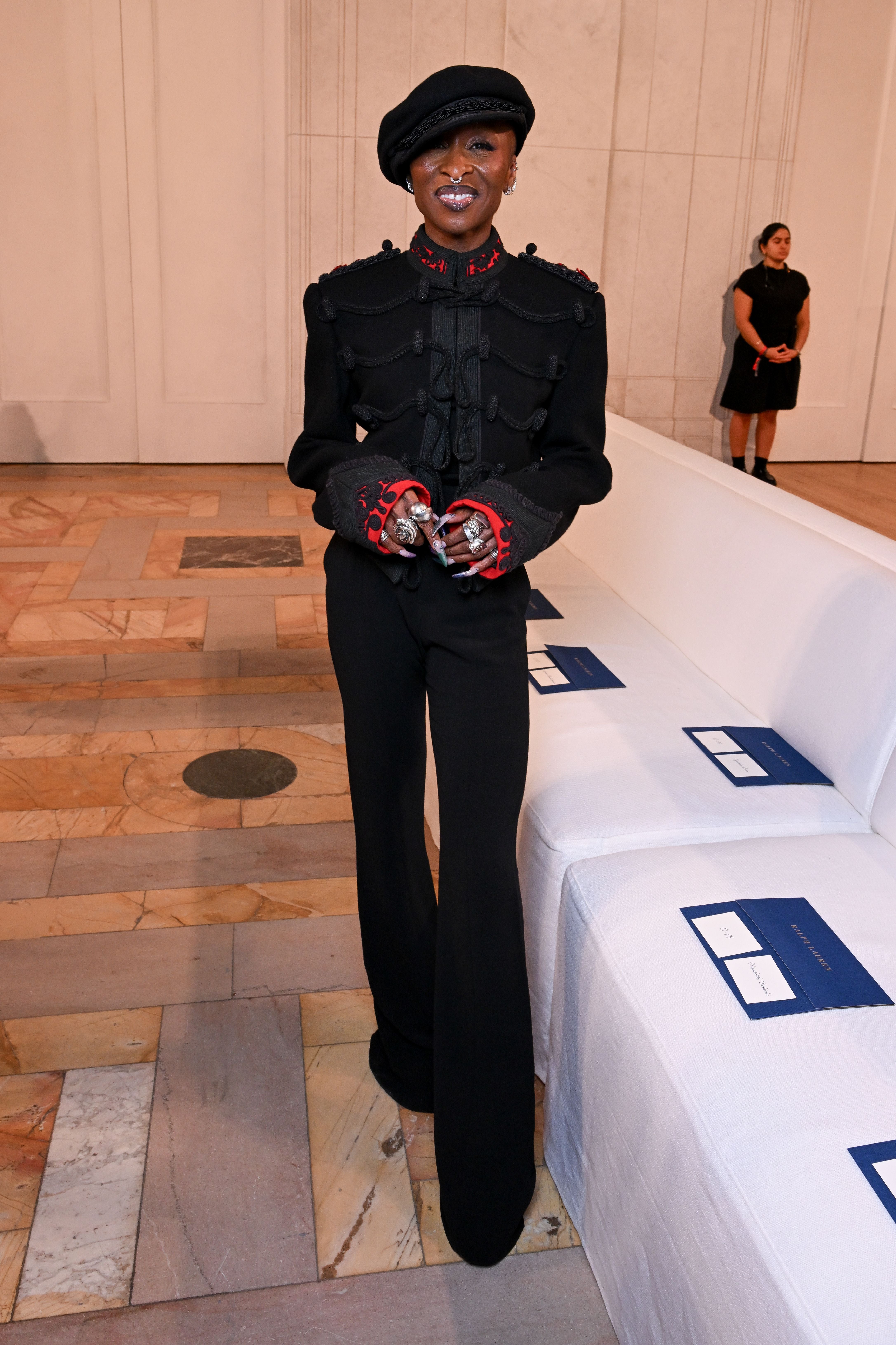 Person mit schwarzem Hut und schwarzem Outfit steht lächelnd neben weißen Sofas in einer Galerie.