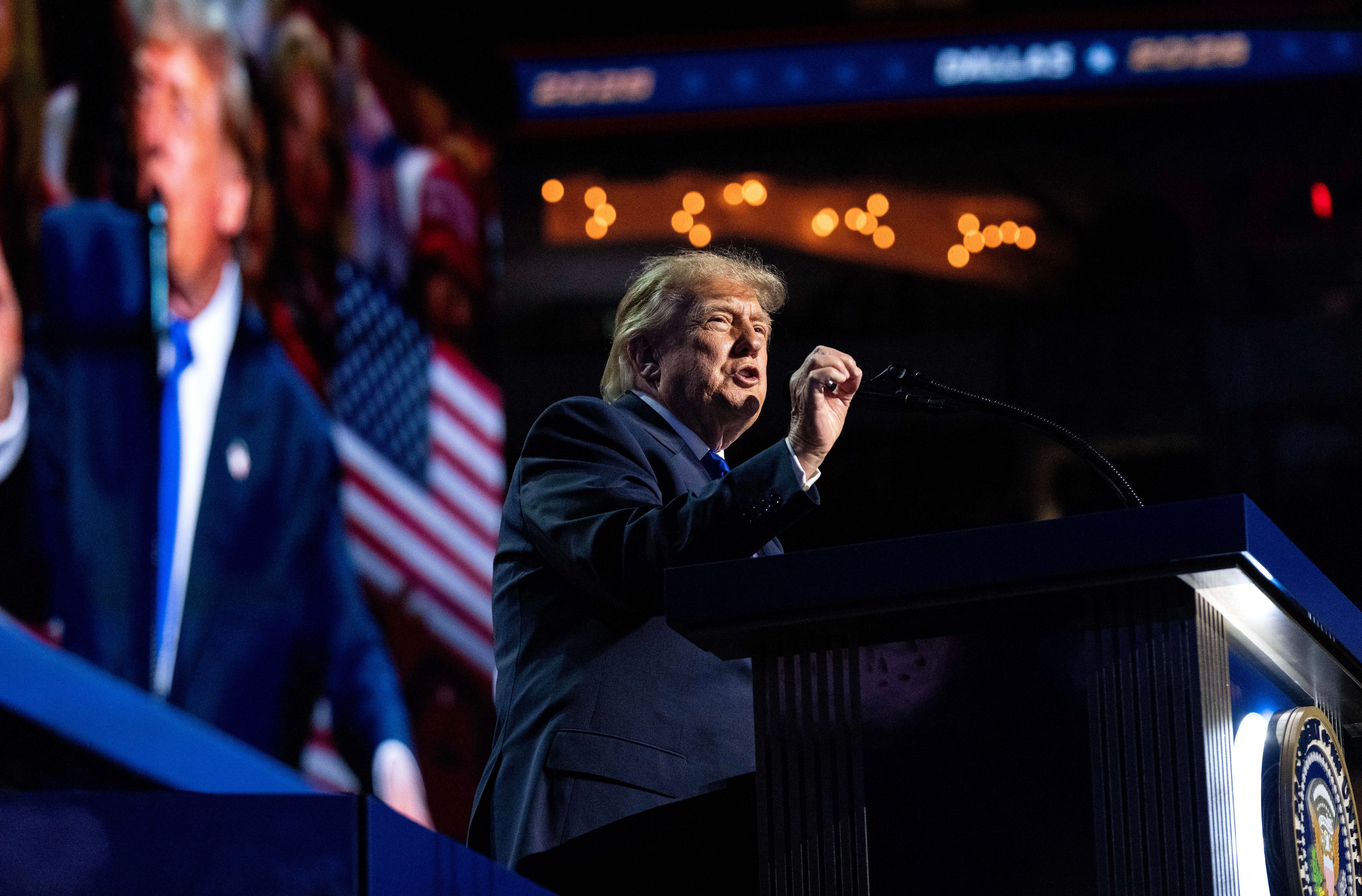 Ein Mann spricht an einem Rednerpult auf einer Bühne, im Hintergrund US-Flagge und Leinwand.