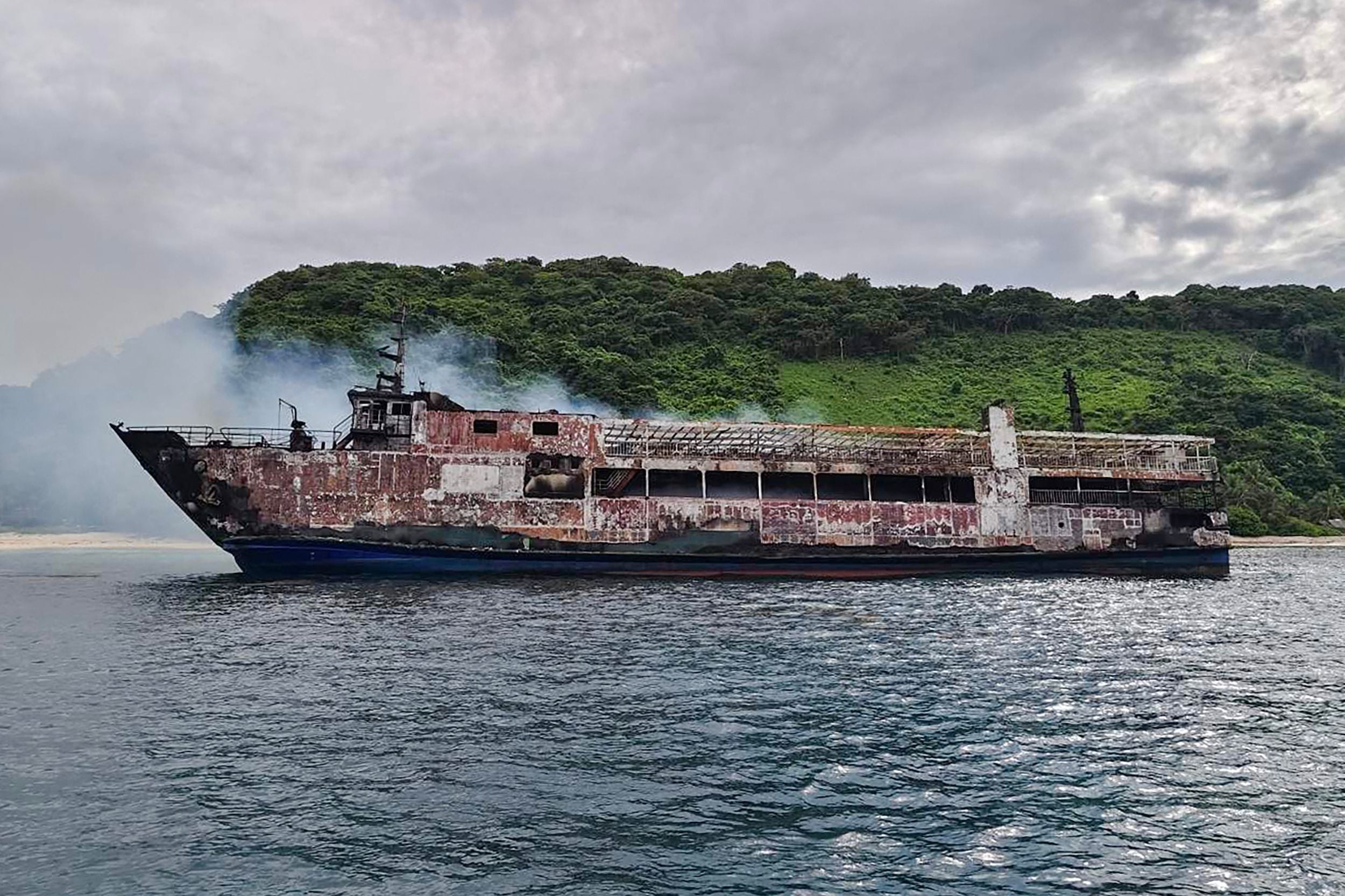 Ein ausgebranntes Fährschiff treibt rauchend vor einer grünen Küste auf dem Wasser.