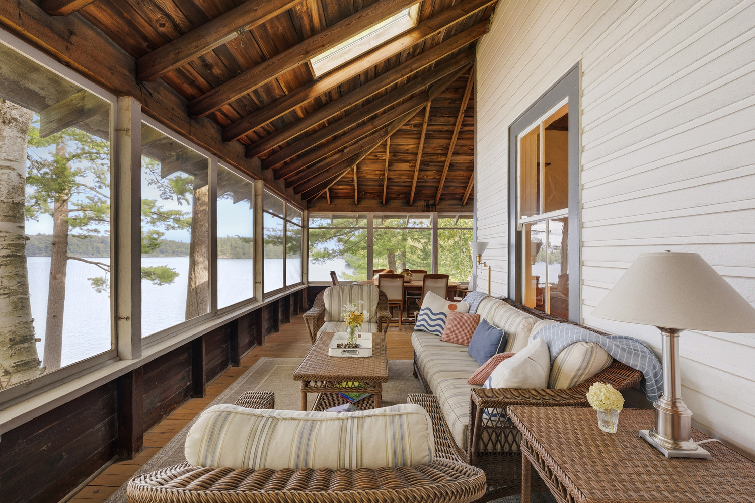 Überdachte Veranda mit geflochtenen Möbeln, Kissen und Blick auf einen See und Bäume.