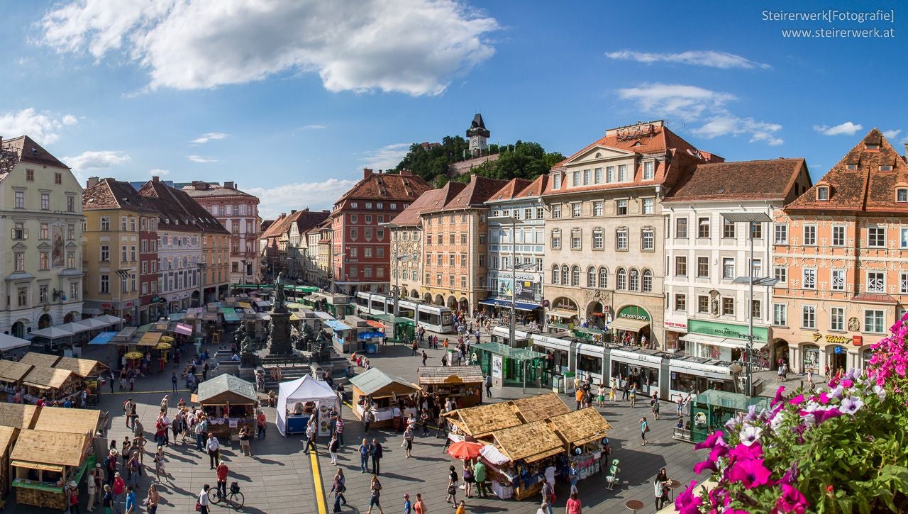 Blick auf den Grazer Hauptplatz mit Marktständen, Straßenbahnen und Menschen bei sonnigem Wetter.