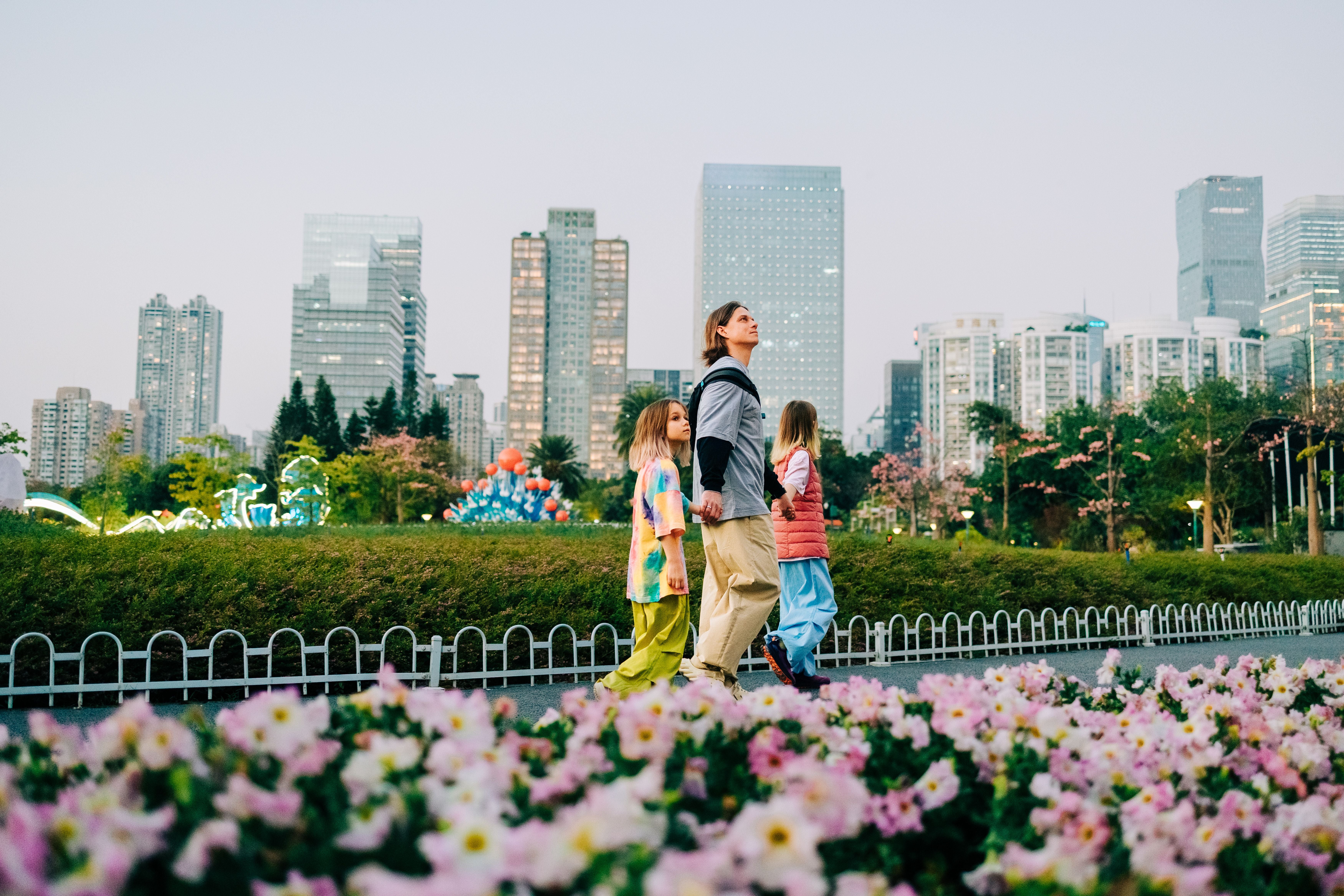 Familie geht Hand in Hand durch einen Stadtpark mit Blumen und Wolkenkratzern im Hintergrund.