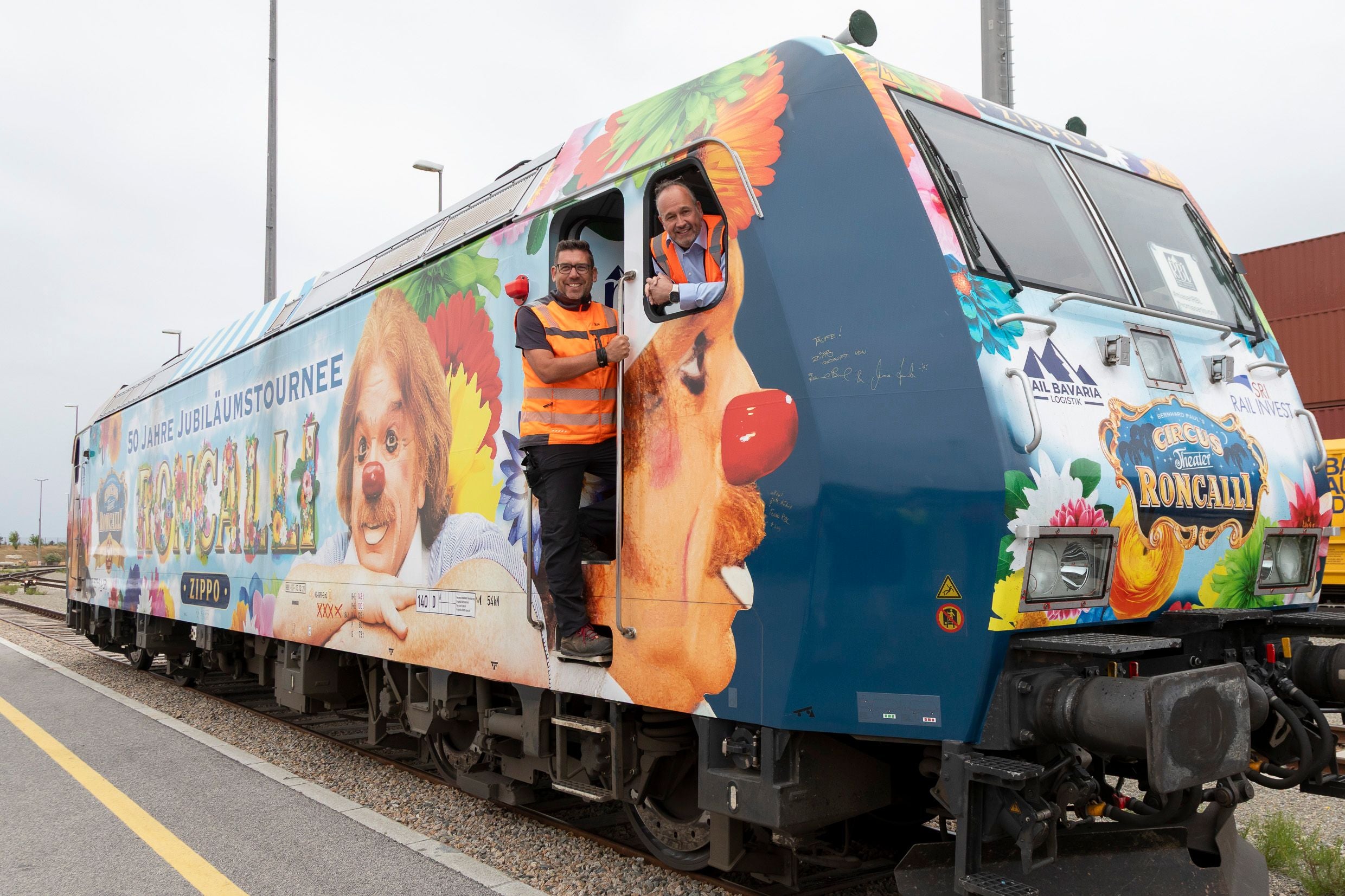 Zwei Männer in Warnwesten stehen an einer bunt bemalten Roncalli-Zuglok mit Clowngesicht.