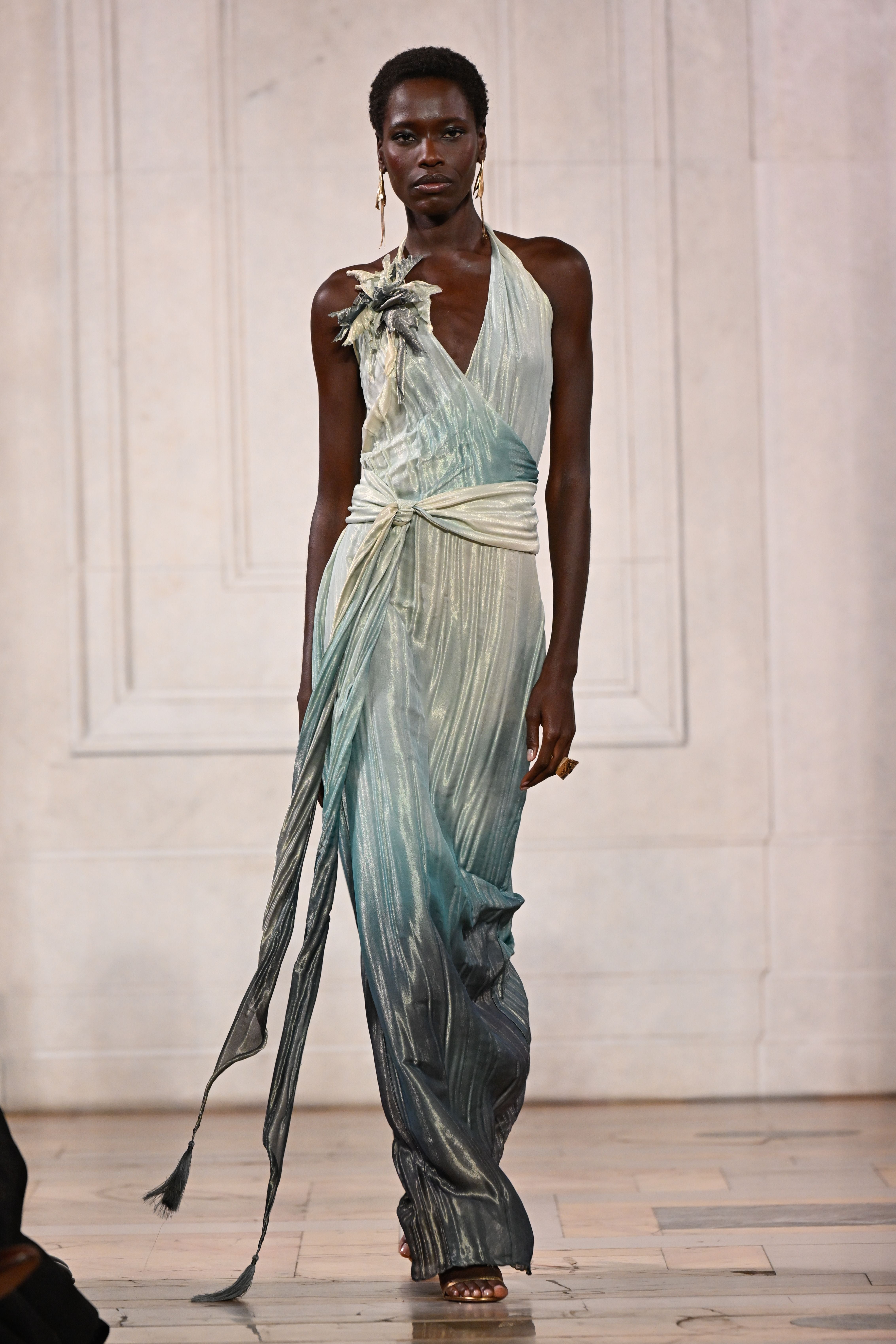 Model präsentiert ein elegantes, fließendes, grün-blaues Kleid auf einem Laufsteg.
