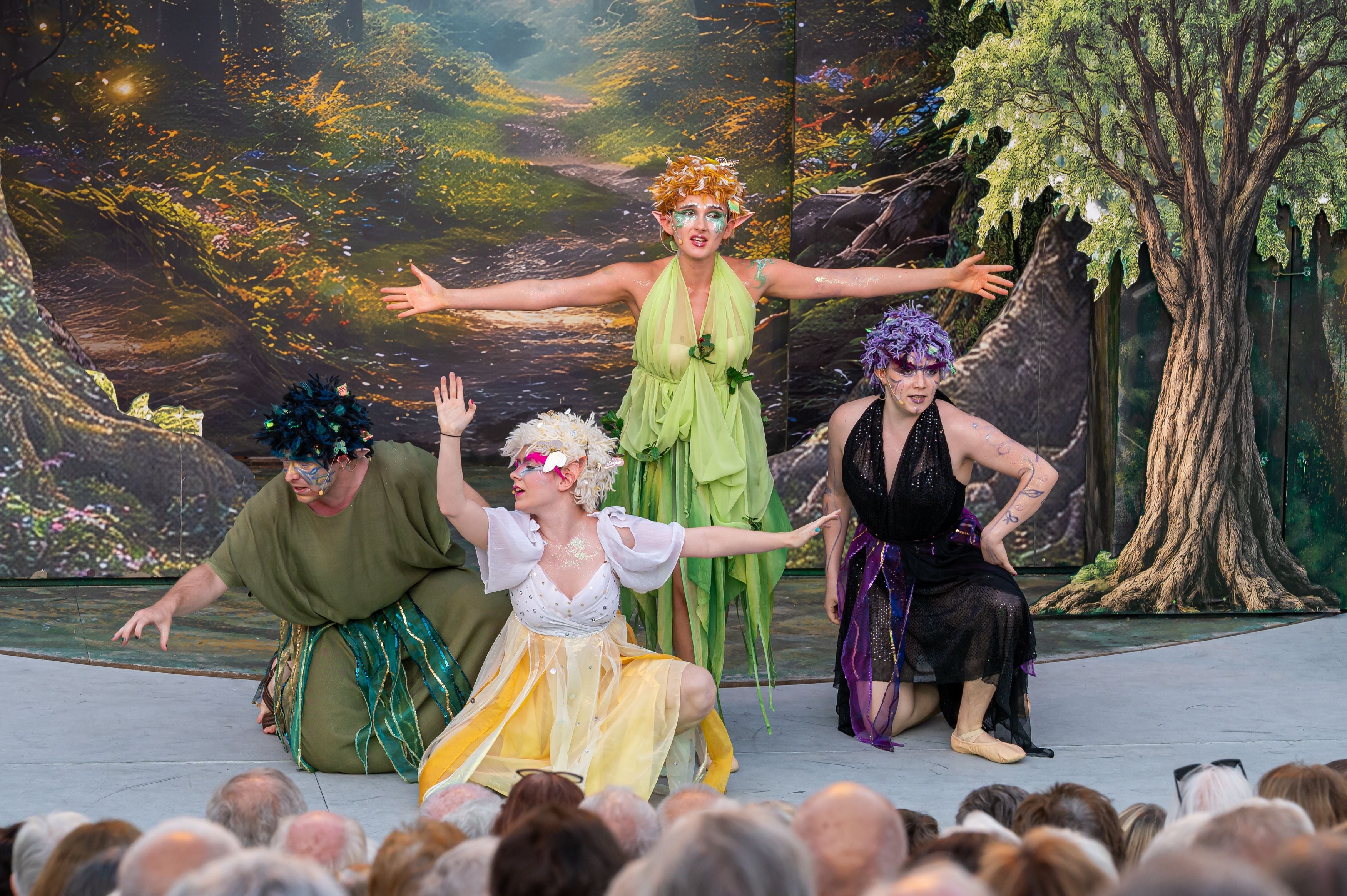Vier Schauspieler in fantasievollen Feenkostümen spielen auf einer Waldbühne vor Publikum.