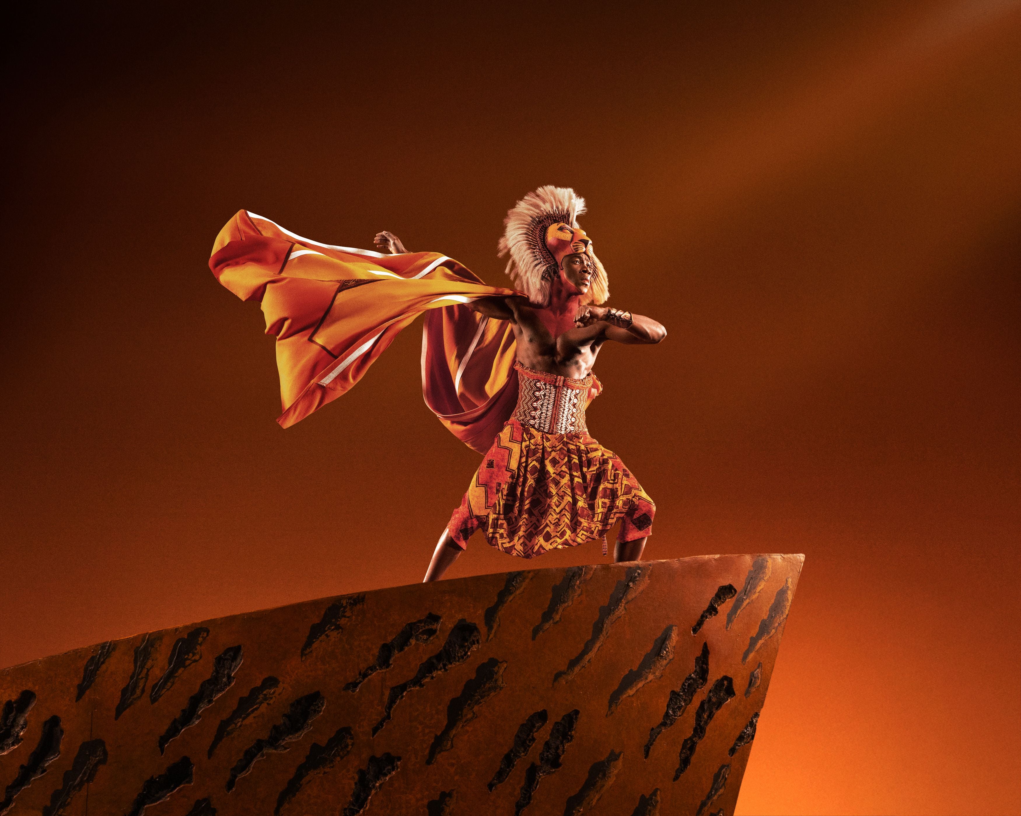 Darsteller als Simba auf dem Königsfelsen vor orangefarbenem Hintergrund, inspiriert von „Der König der Löwen“.