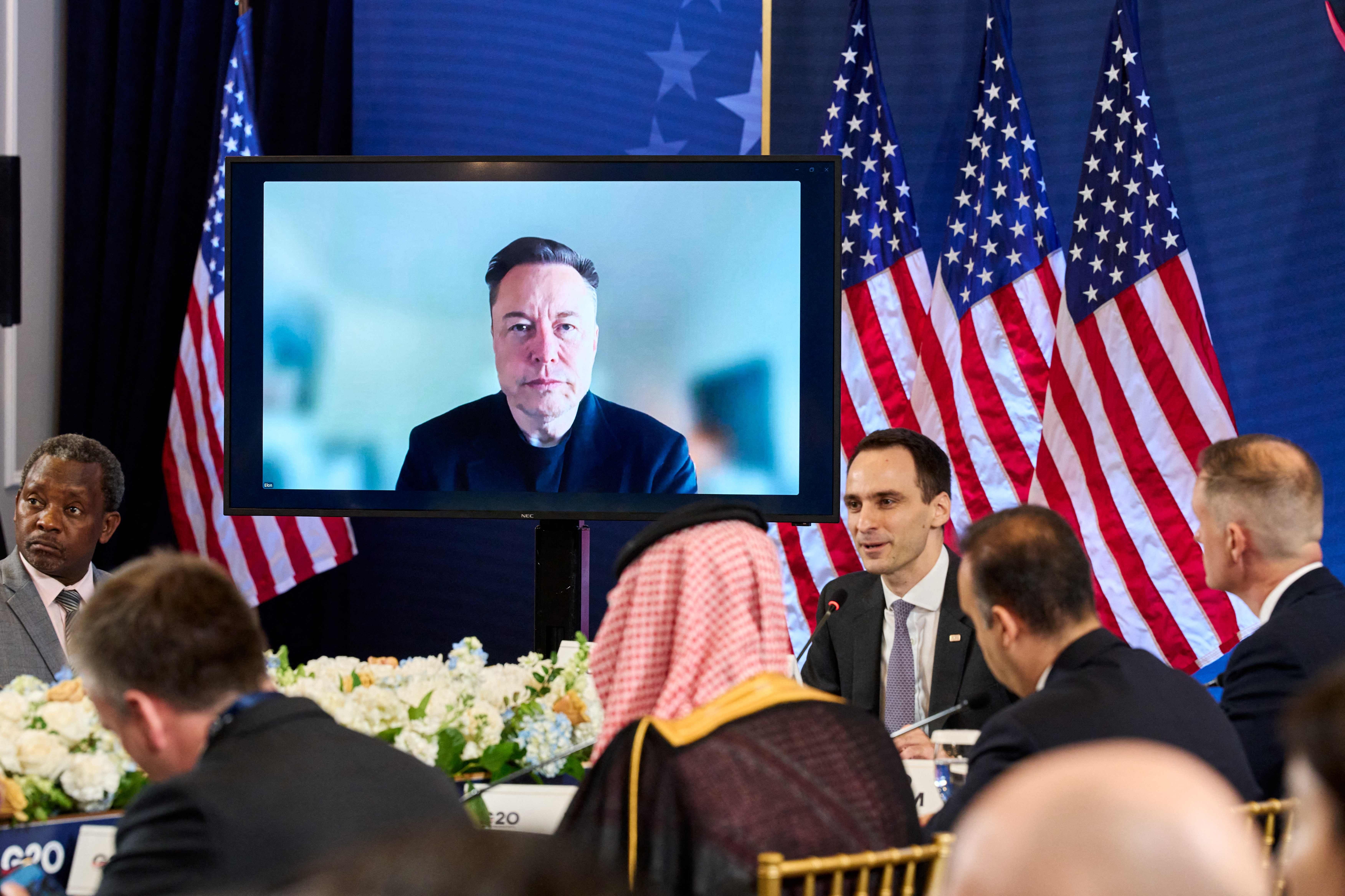 Elon Musk nimmt per Video an einem G20-Gipfel mit US-Flaggen im Hintergrund teil.