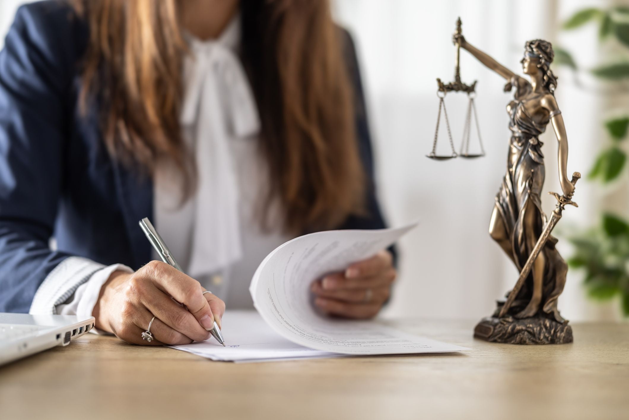 Frau unterschreibt Dokumente im Büro, daneben eine Justitia-Statue und ein Laptop.