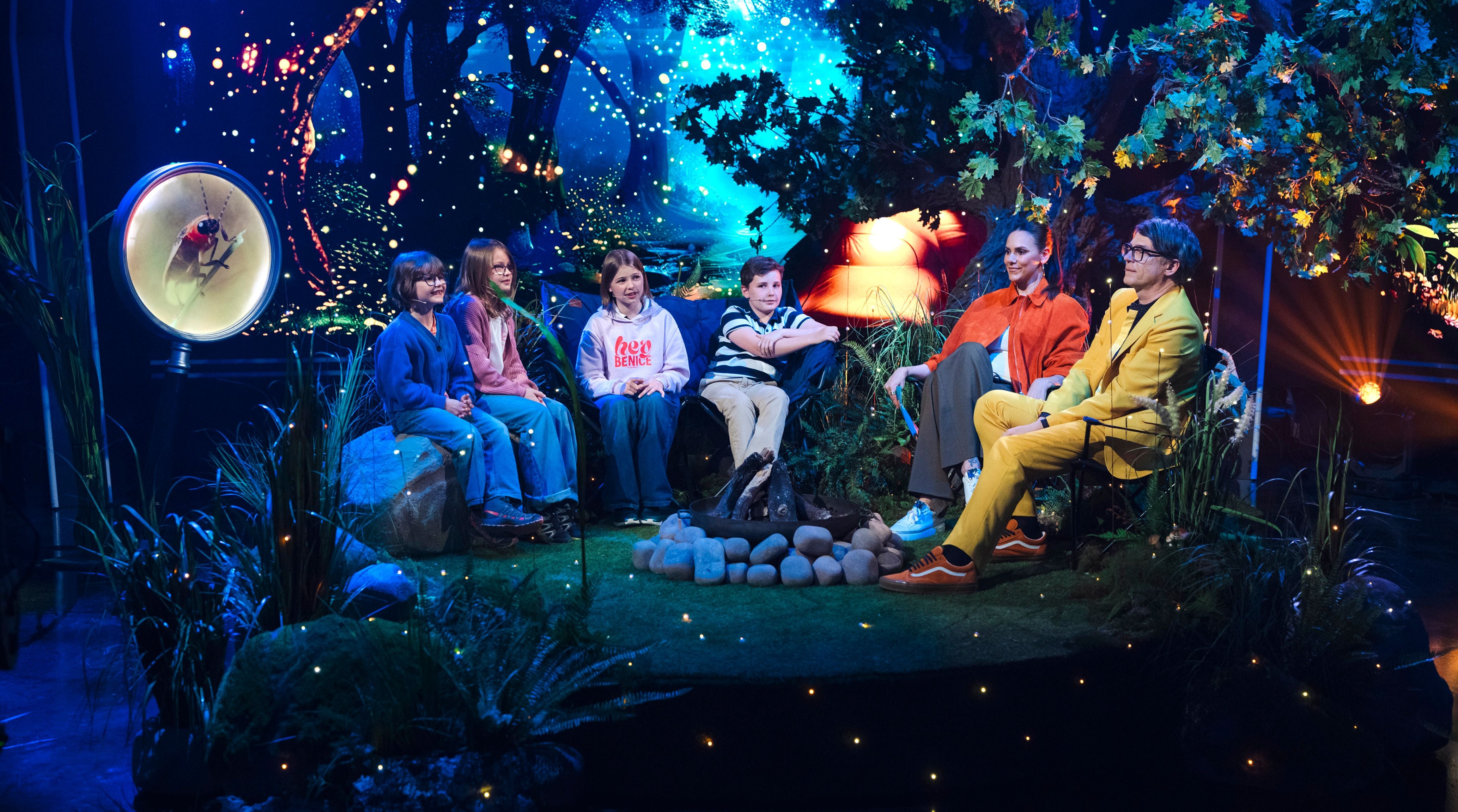 Mehrere Kinder und zwei Erwachsene sitzen in einer Wald-Kulisse um ein Lagerfeuer im Fernsehstudio.