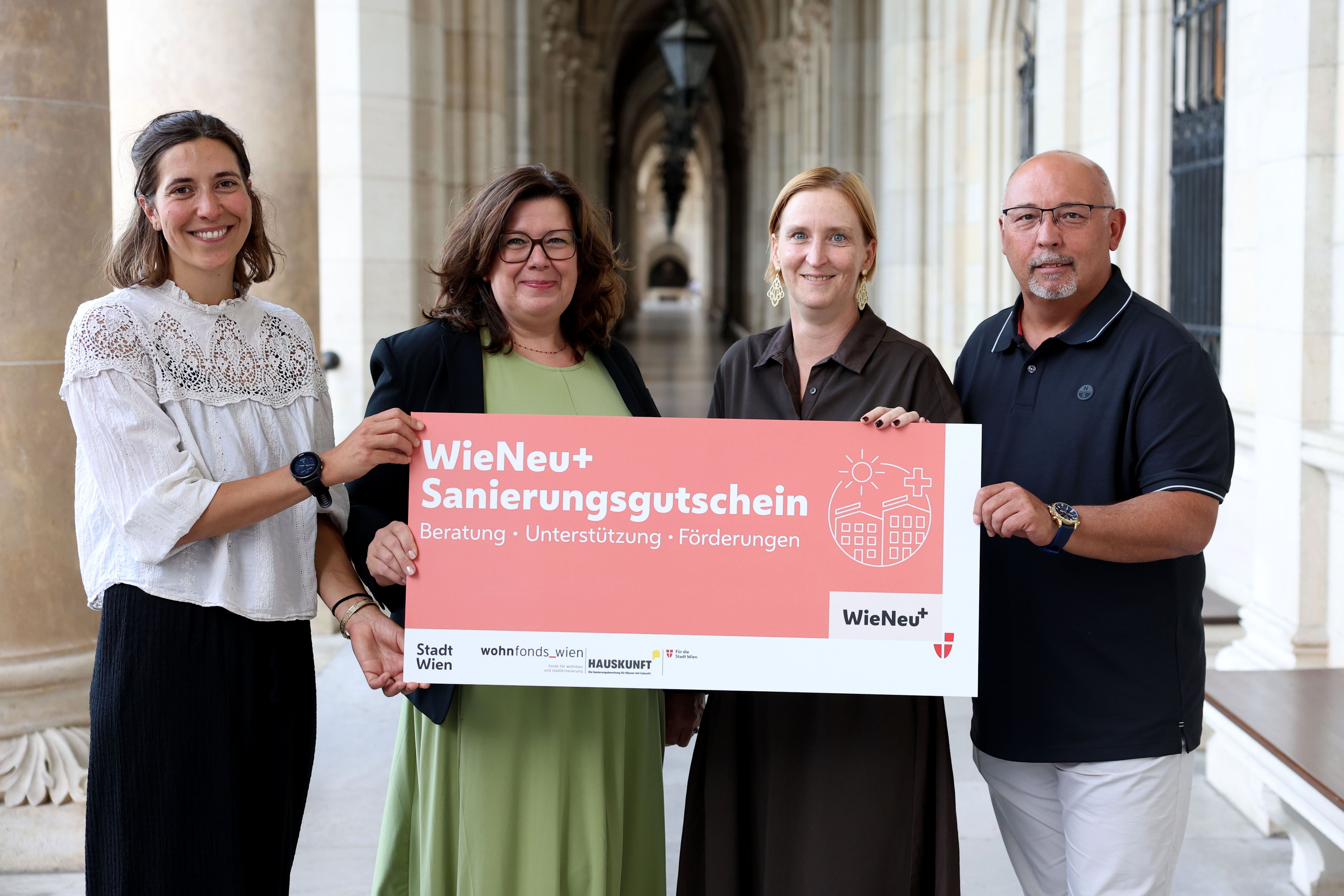 Vier Personen halten ein Schild mit der Aufschrift "WieNeu+ Sanierungsgutschein" in einem Säulengang.