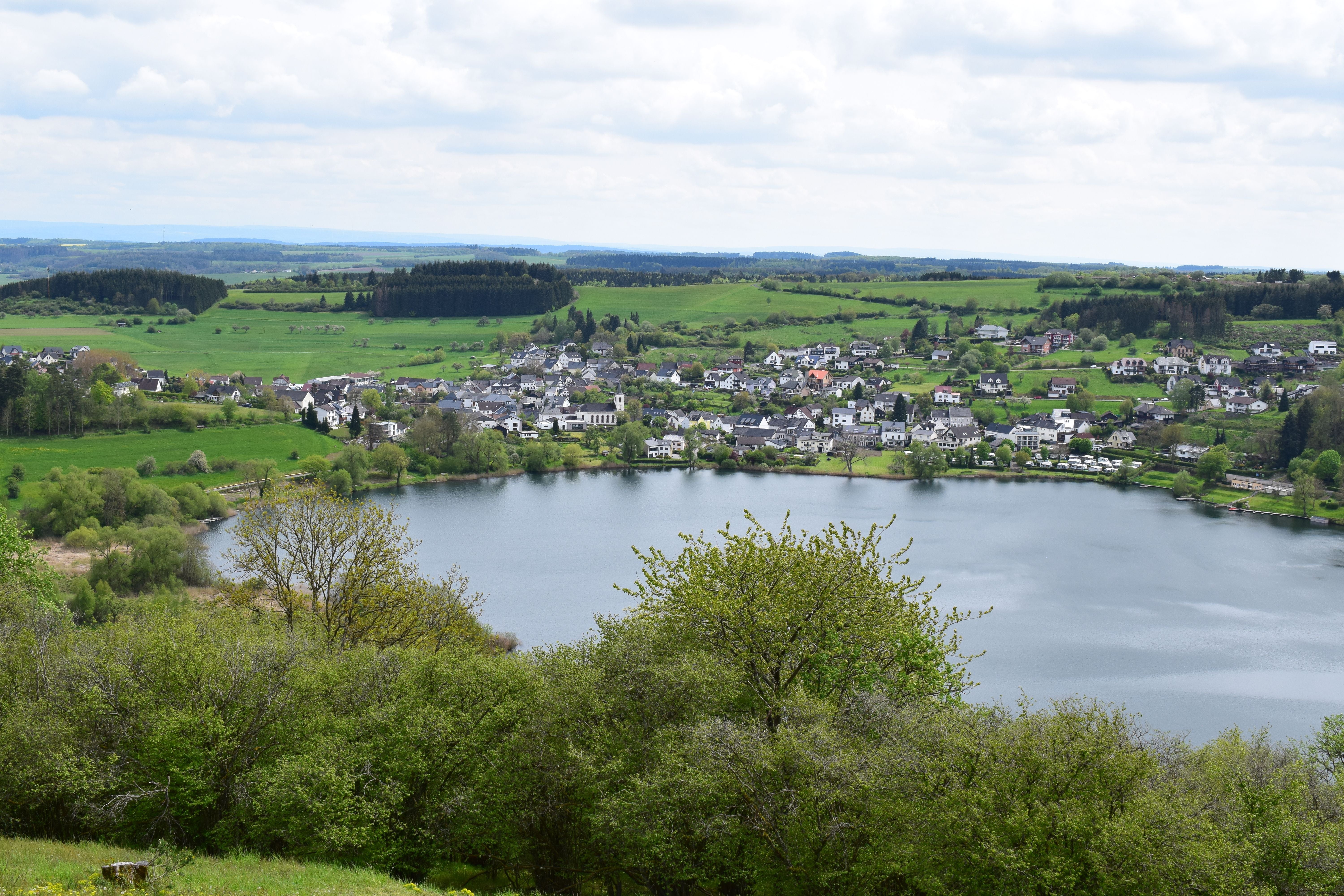 Blick auf das Schalkenmehrener Maar mit dem Ort Schalkenmehren und grüner Frühlingslandschaft.