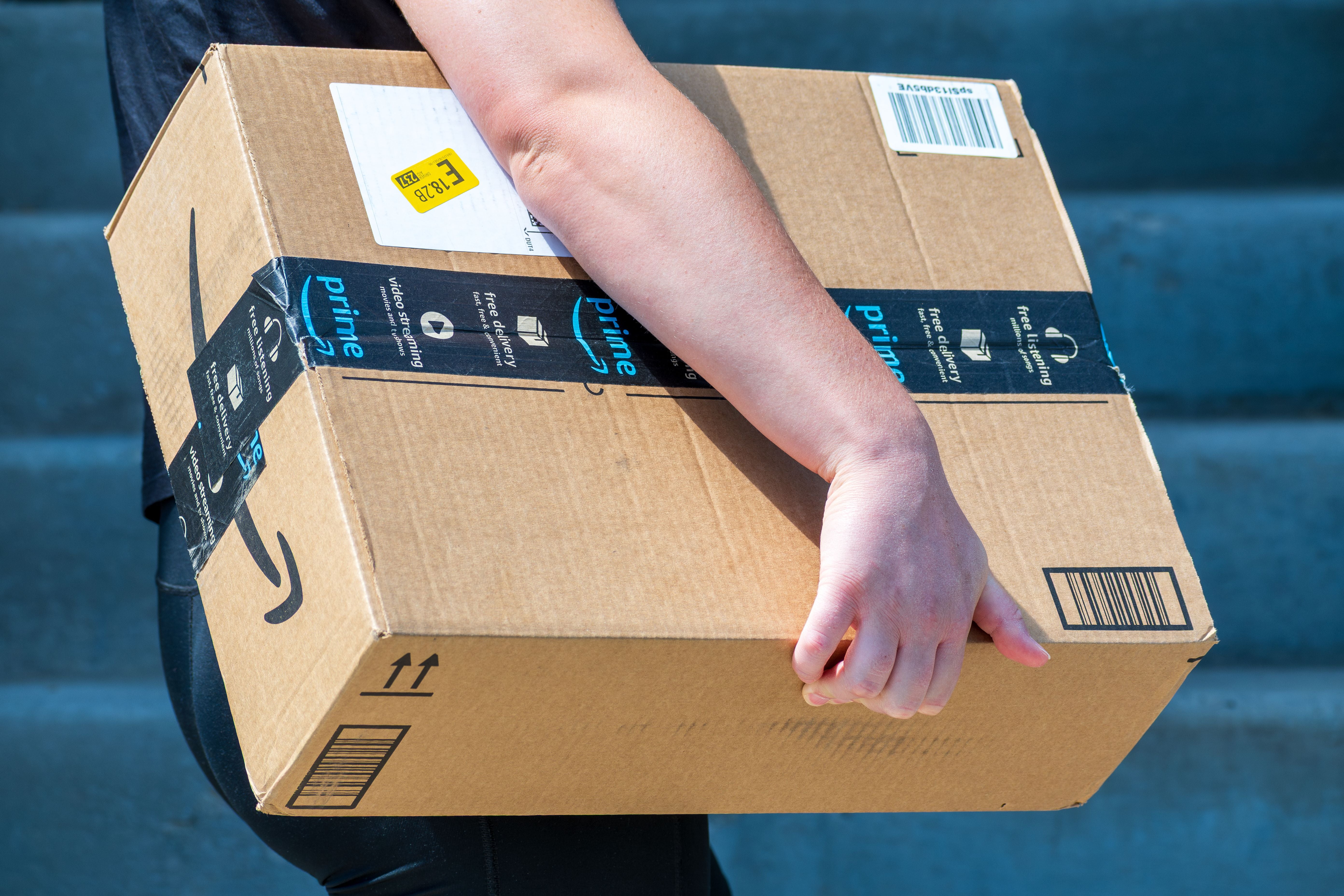 Person trägt ein großes Amazon-Paket zu einer Haustür.