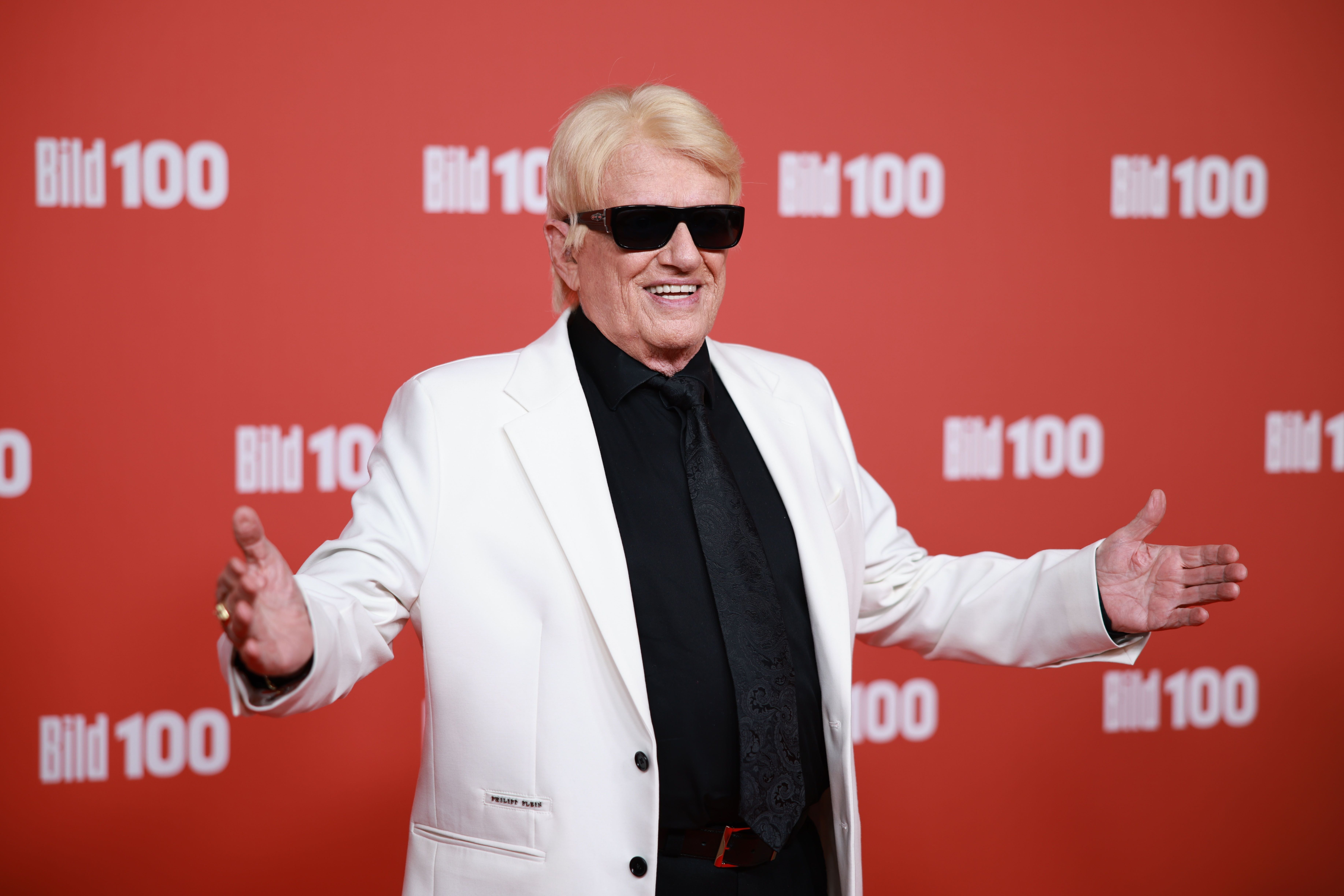 Ein Mann mit blondem Haar, weißem Anzug und Sonnenbrille posiert vor rotem "Bild 100"-Hintergrund.