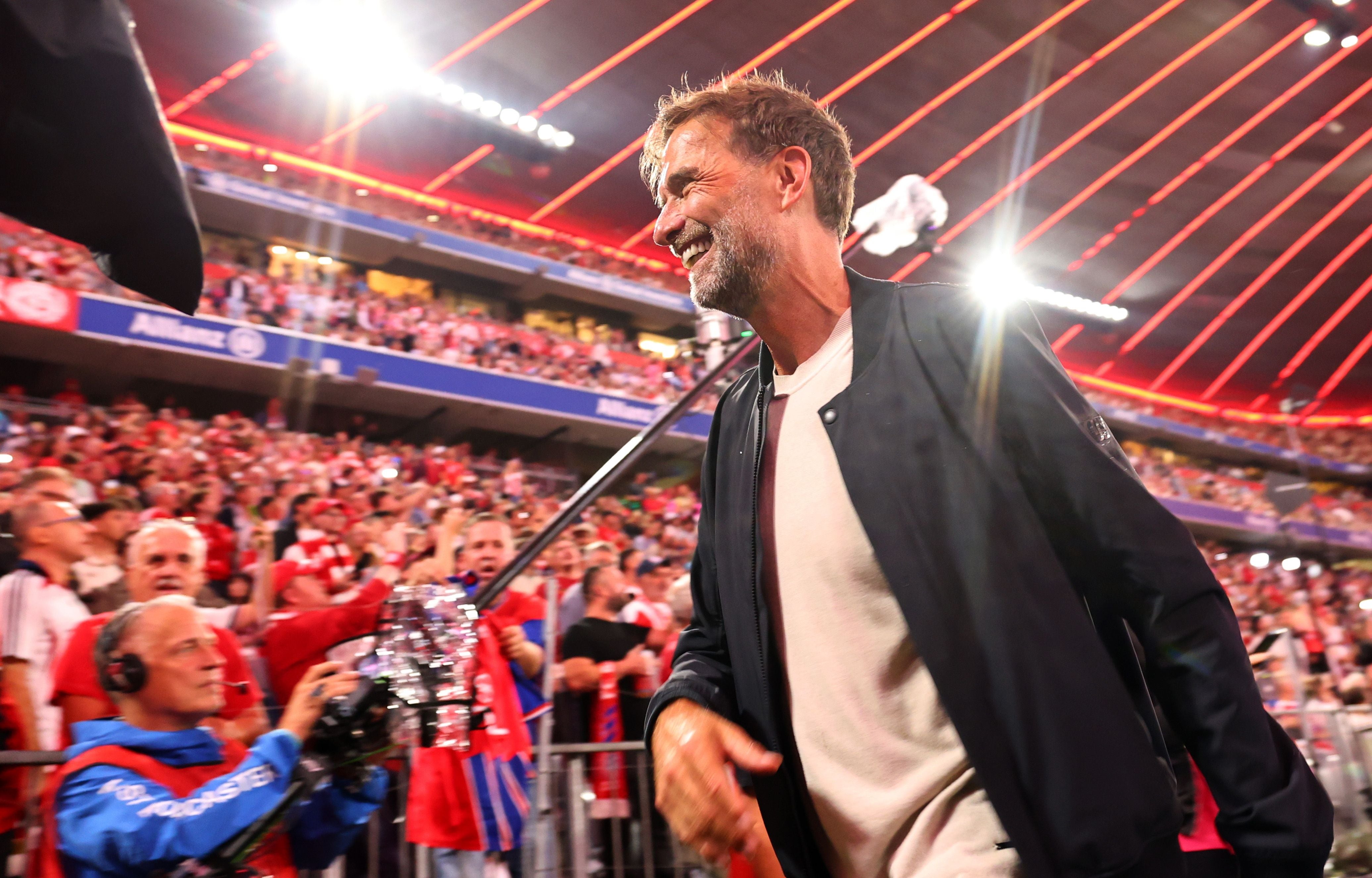 Ein lächelnder Mann im Stadion mit jubelnden Fans im Hintergrund bei einem Fußballspiel.