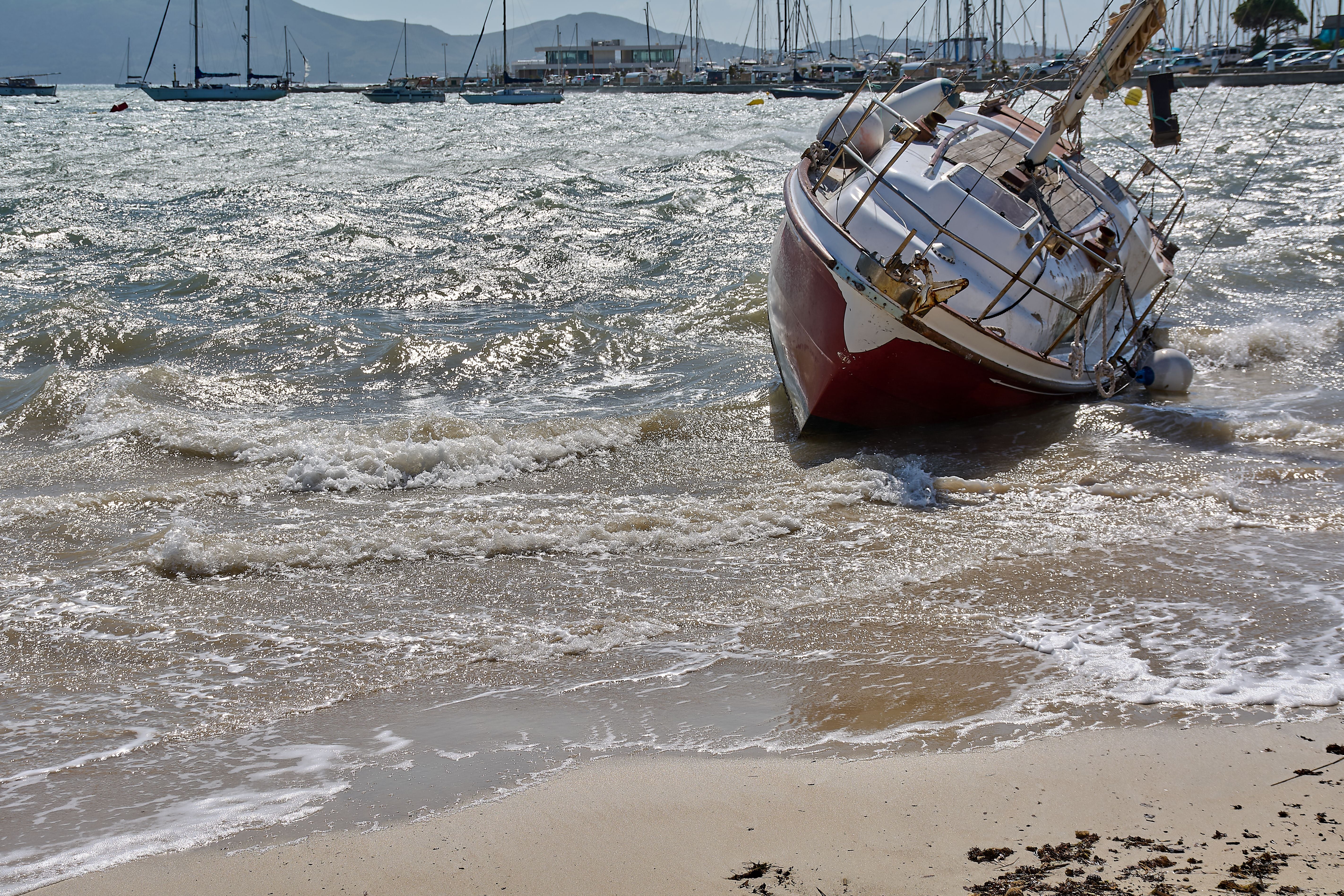 Gescheuertes weiß-rotes Segelboot liegt am Sandstrand von Puerto Pollenca, Mallorca, bei Wellengang.