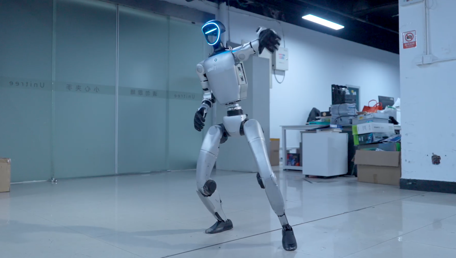 Ein humanoider Roboter steht in einem modernen, etwas unordentlichen Büro und hebt einen Arm.