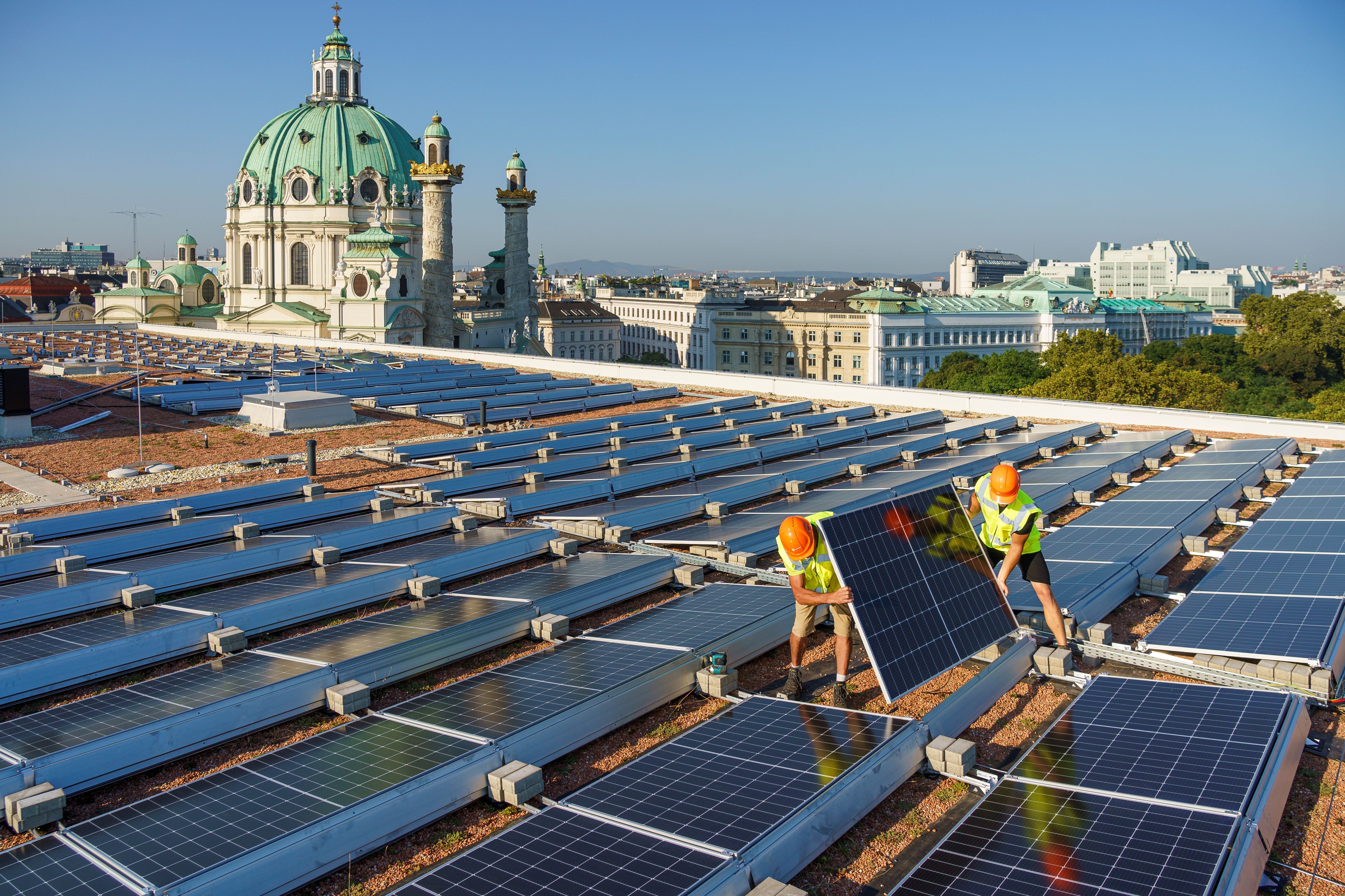 Zwei Arbeiter installieren Solarpanels auf einem Dach mit Stadt und Kirche im Hintergrund.