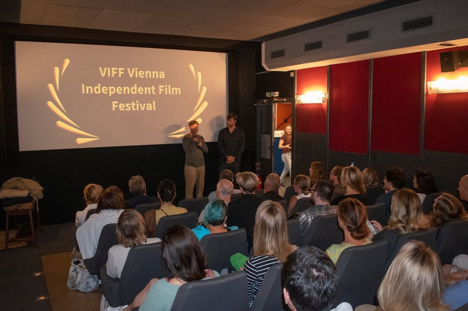 Menschen sitzen in einem Kinosaal beim Vienna Independent Film Festival, zwei Männer stehen vorne.