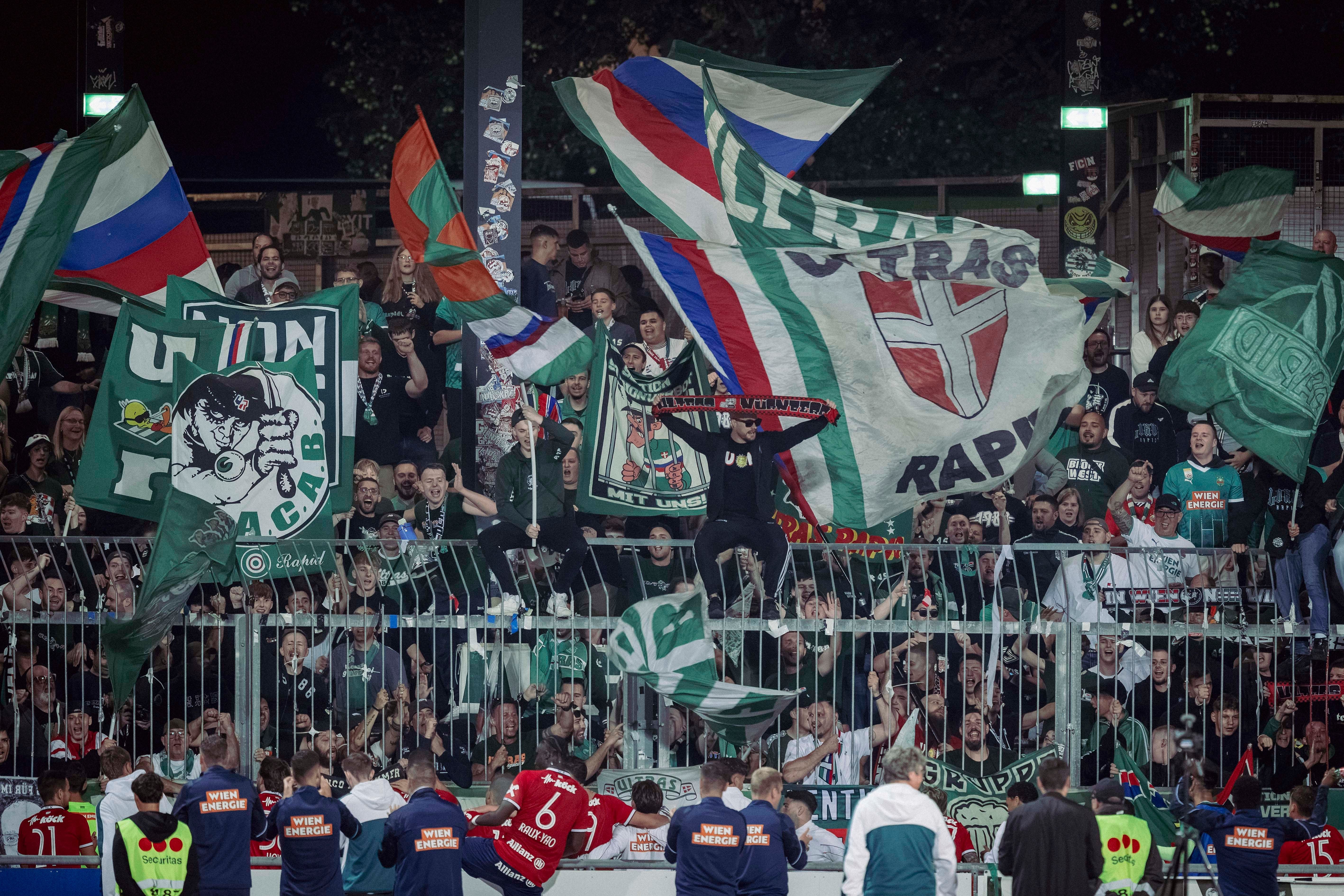 Rapid Wien-Fans feiern im Stadion mit grünen Fahnen und Transparenten hinter dem Spielfeldzaun.