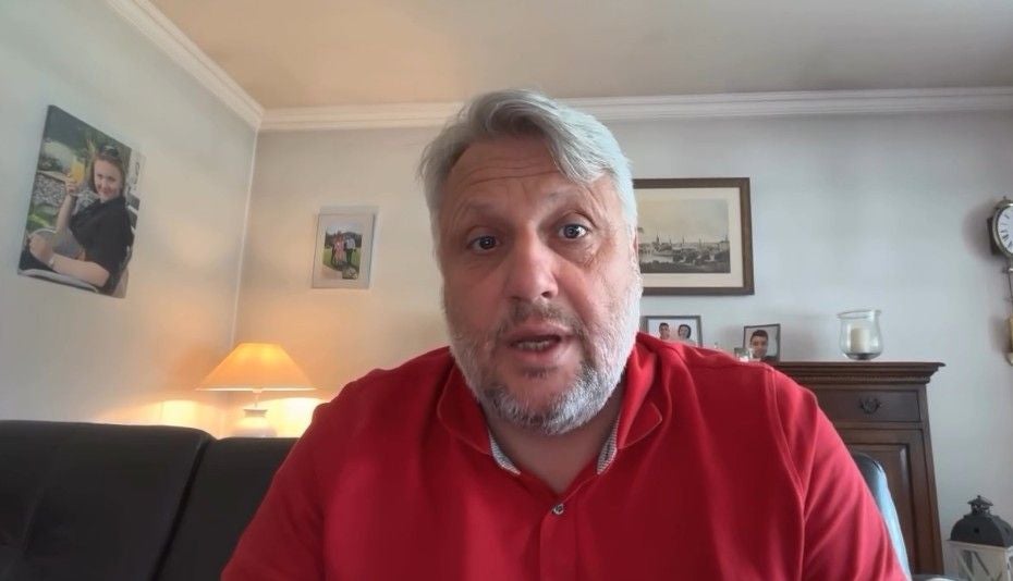 Mann mit grauen Haaren und rotem Poloshirt sitzt in einem Wohnzimmer und blickt in die Kamera.