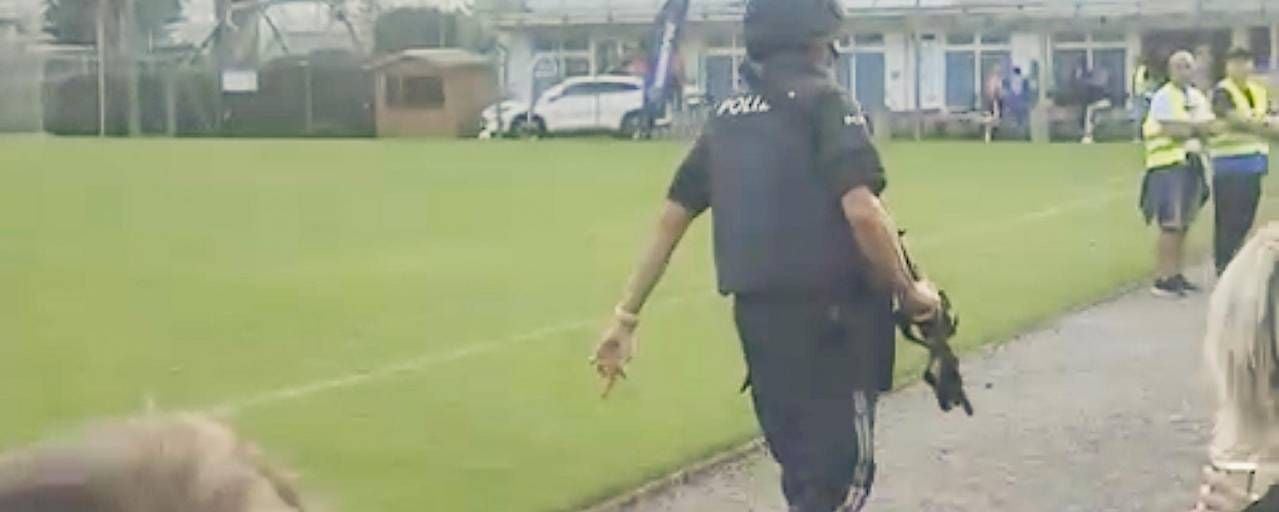 Bewaffneter Polizist geht auf einem Sportplatz, Menschen stehen im Hintergrund.