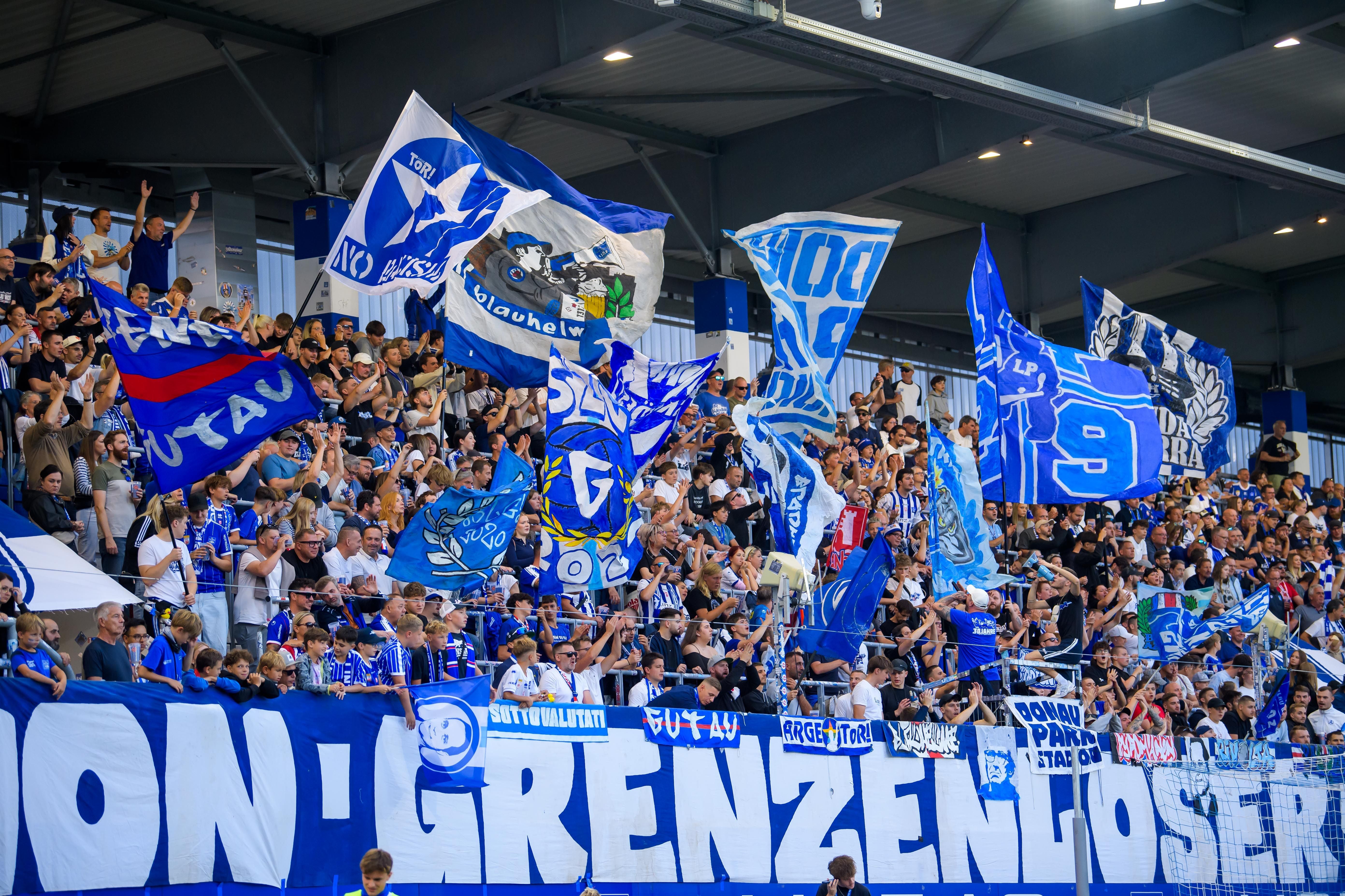 Fans von Blau Weiss Linz jubeln im Stadion mit vielen blauen und weißen Fahnen und Bannern.