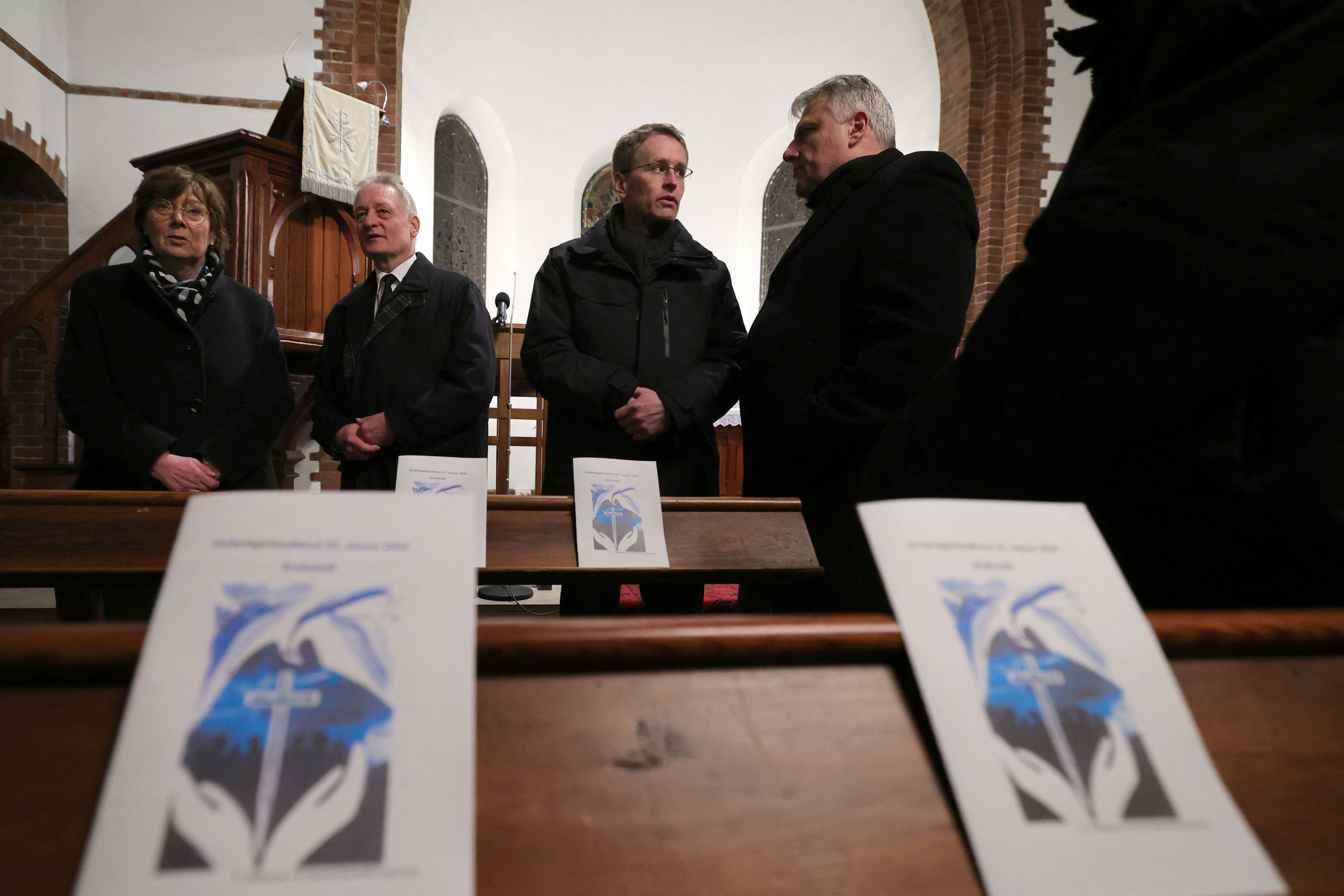 Vier Menschen stehen in einer Kirche bei einer Gedenkfeier, vorn liegen Gedenkprogramme auf den Bänken.