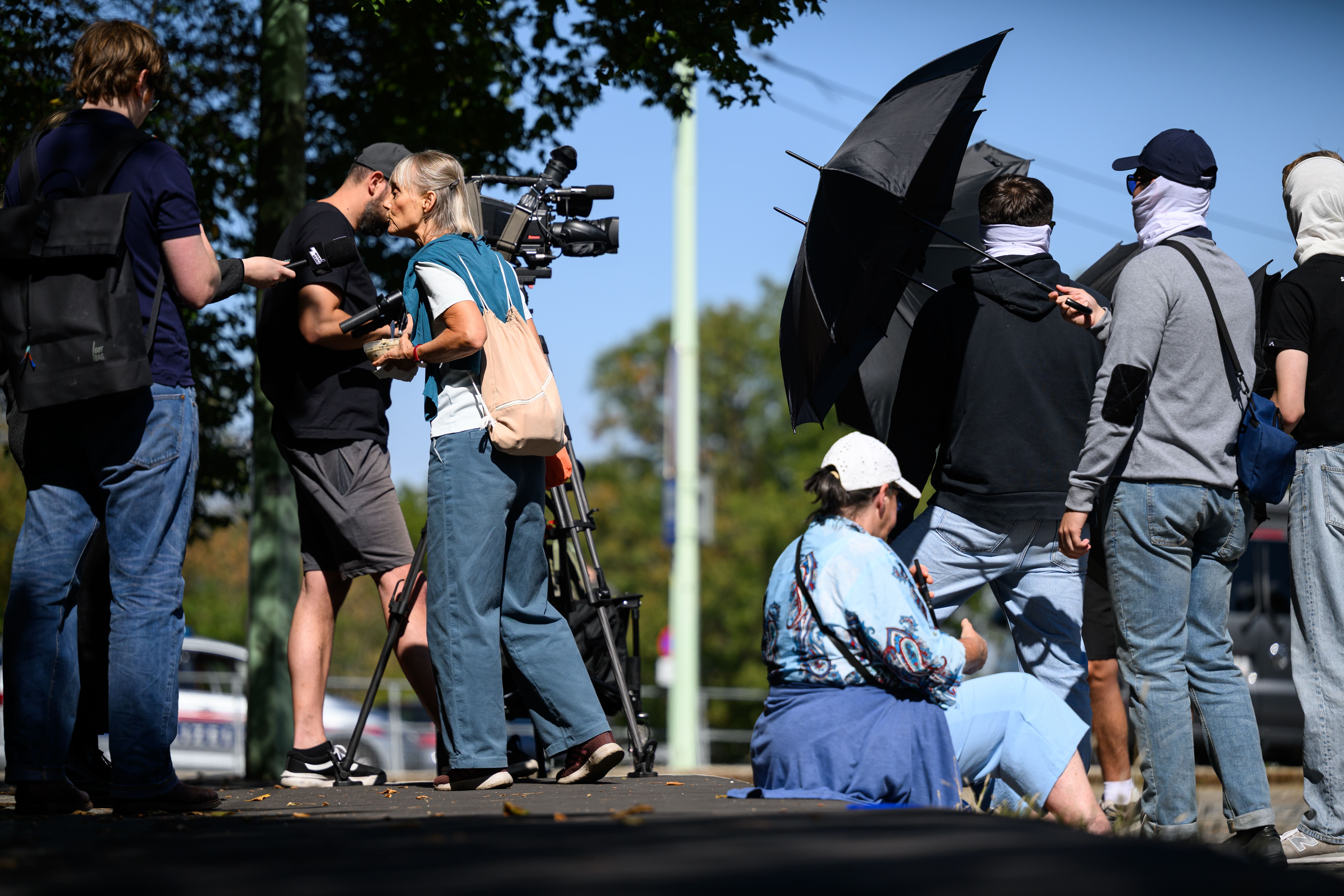 Mehrere Personen mit Schirmen und vermummten Gesichtern versammeln sich im Freien, teils mit Medien.
