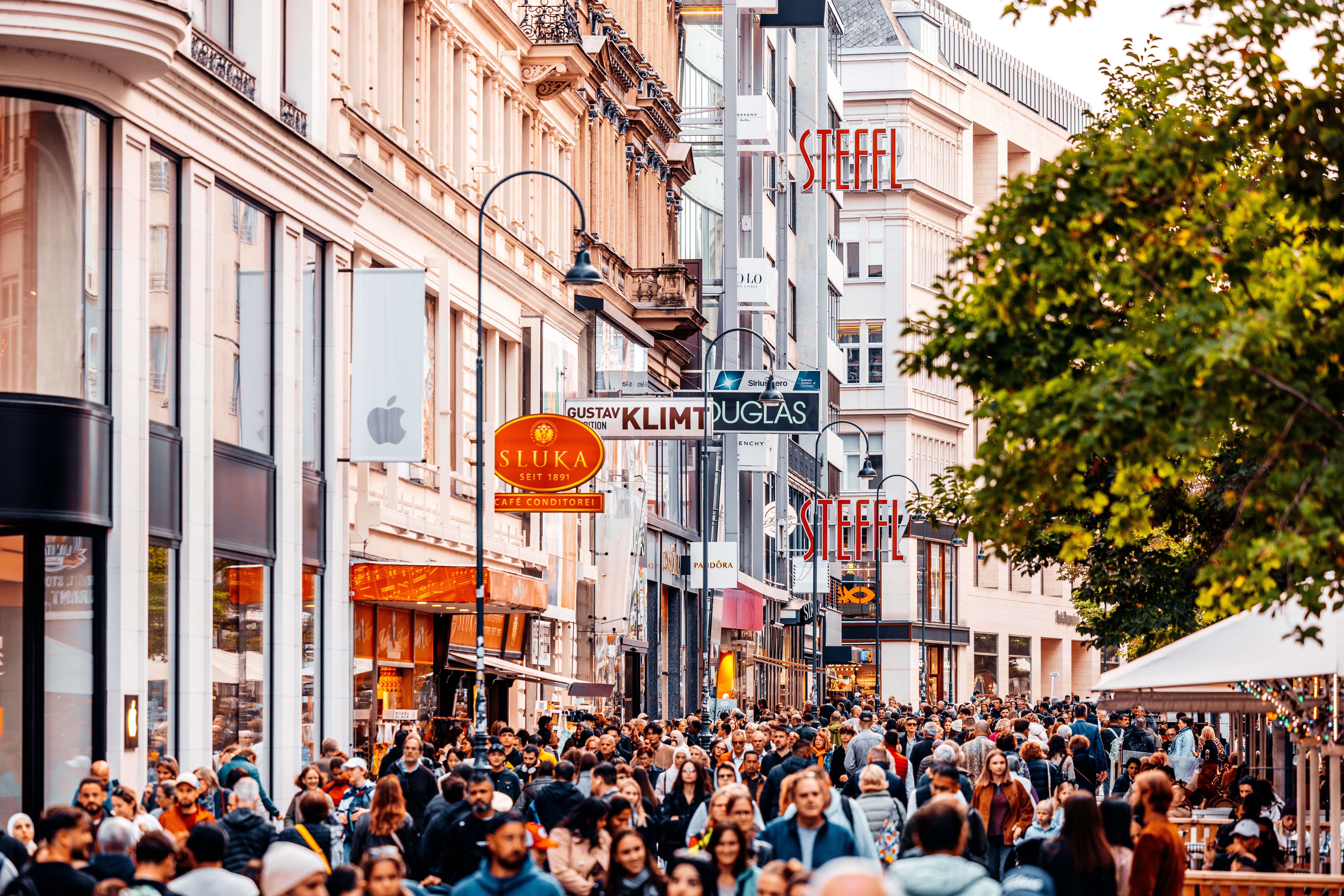 Belebte Einkaufsstraße in Wien mit vielen Menschen und verschiedenen Geschäftsschildern.