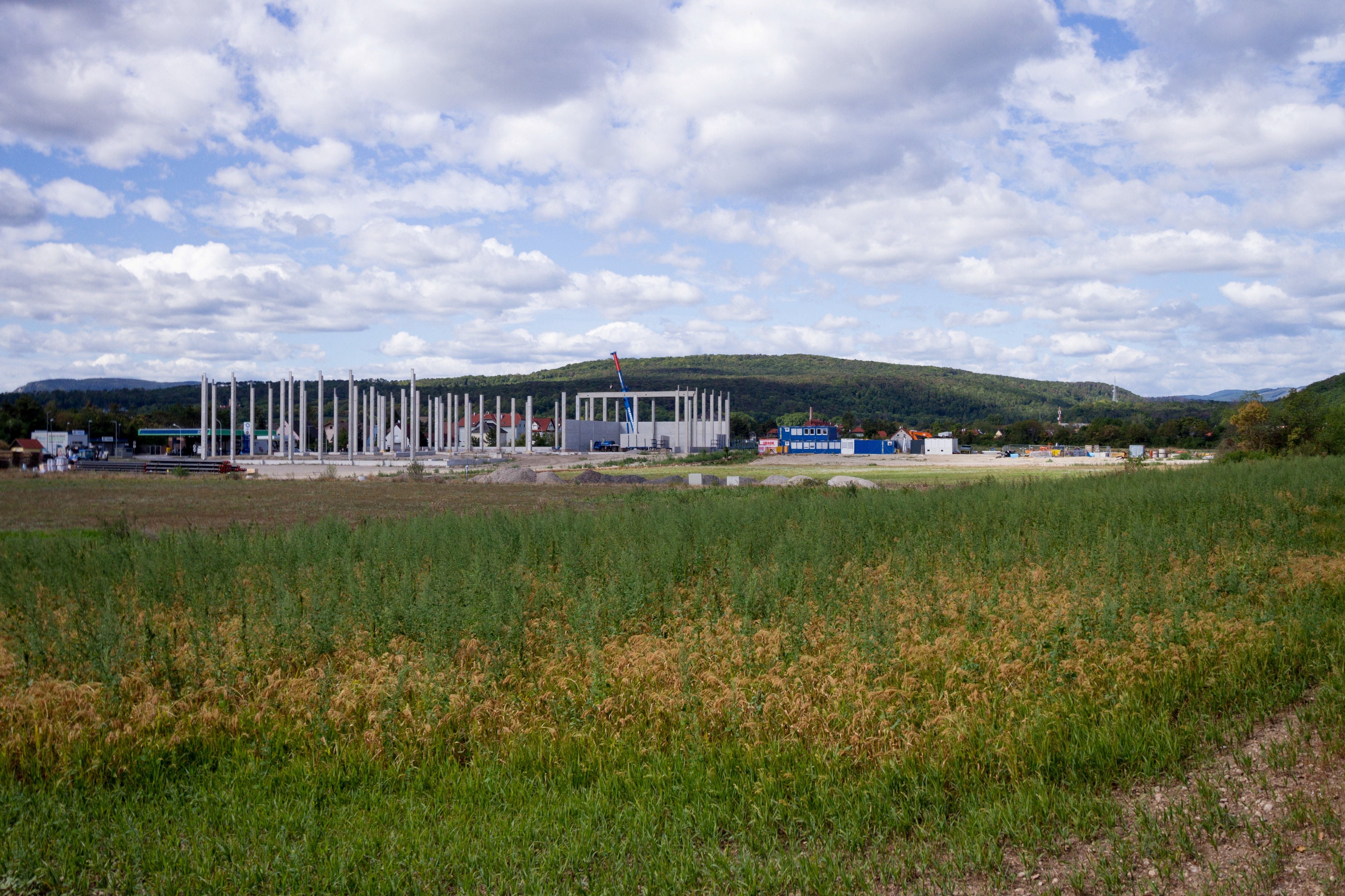 Blick auf eine Baustelle mit Betonsäulen und Wiese, im Hintergrund bewaldete Hügel und Wolken.