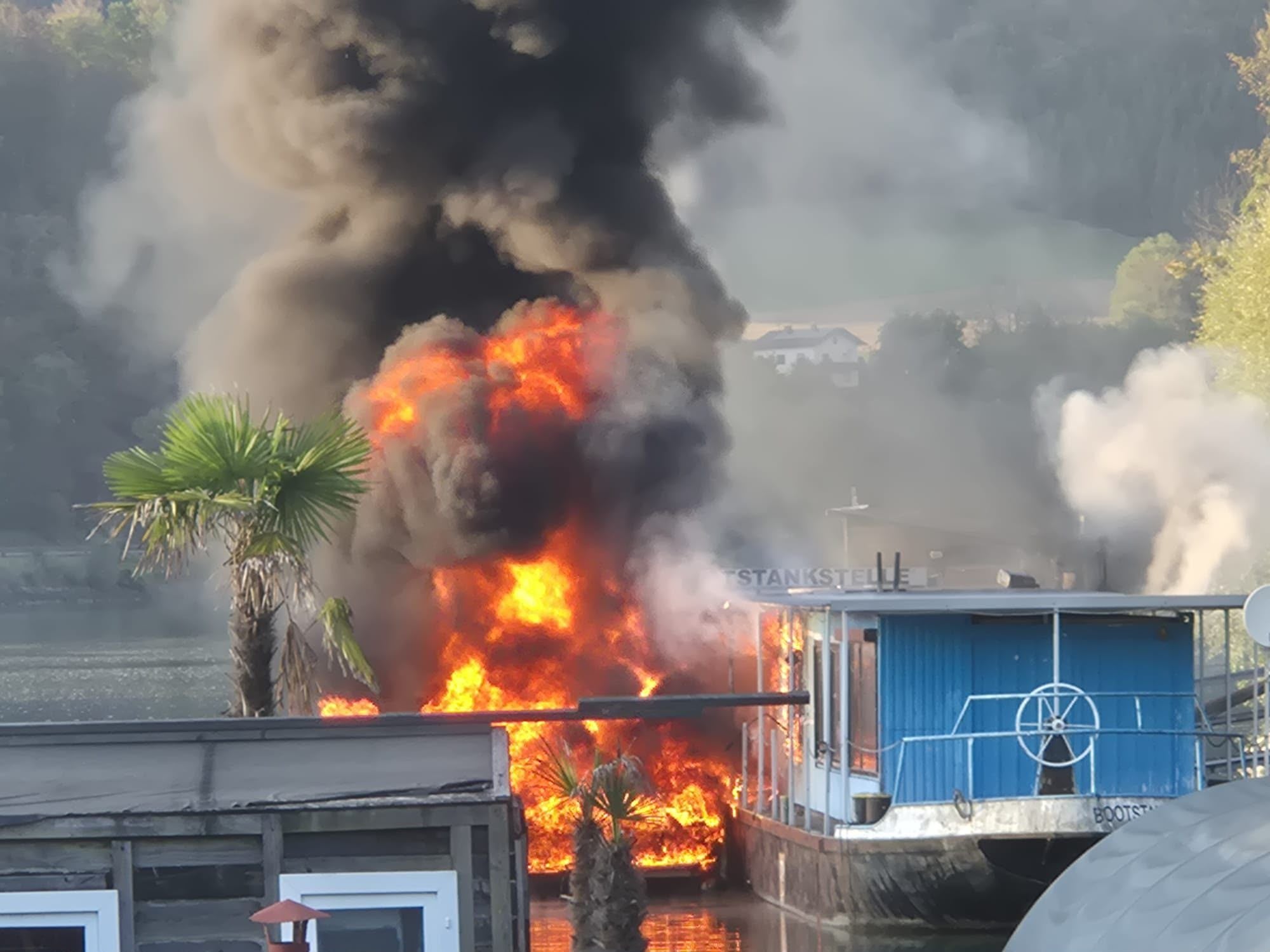 Ein Boot steht an einer Bootstankstelle am Ufer in Flammen, dicker Rauch steigt auf.
