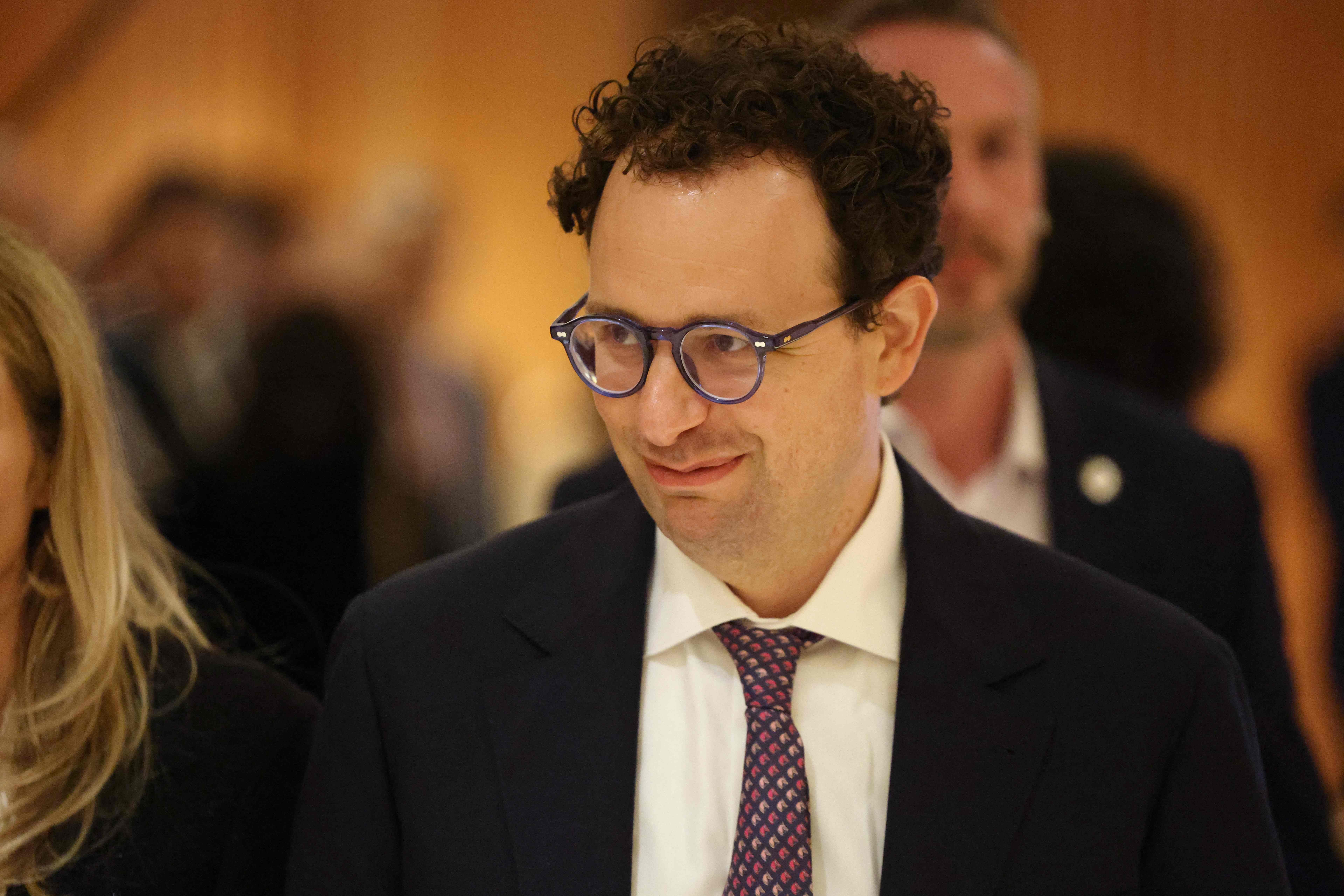Ein Mann mit lockigen Haaren und Brille trägt Anzug und Krawatte, Hintergrund unscharf.