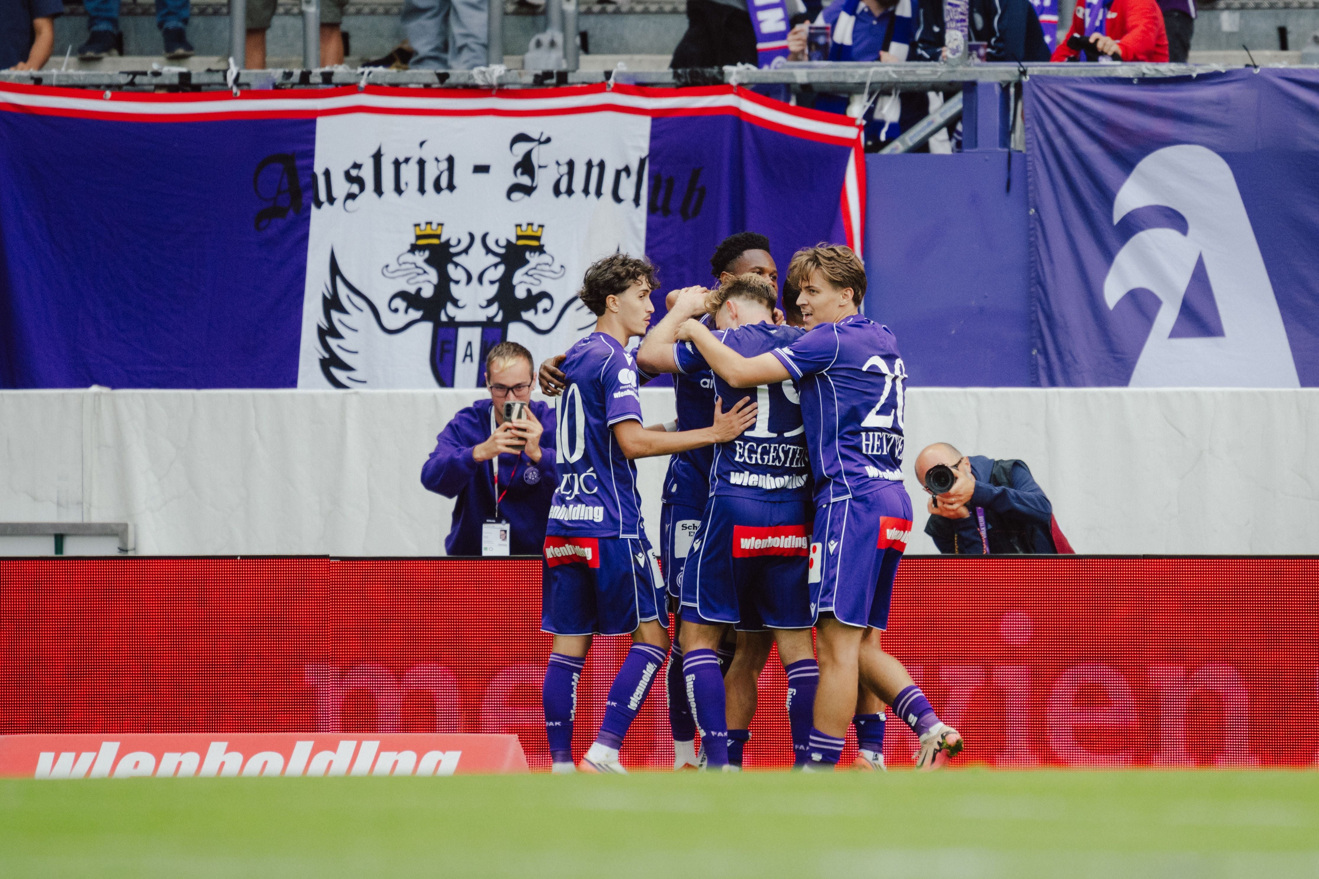 Spieler von Austria Wien jubeln gemeinsam vor einem Austria-Fanclub-Banner im Stadion.