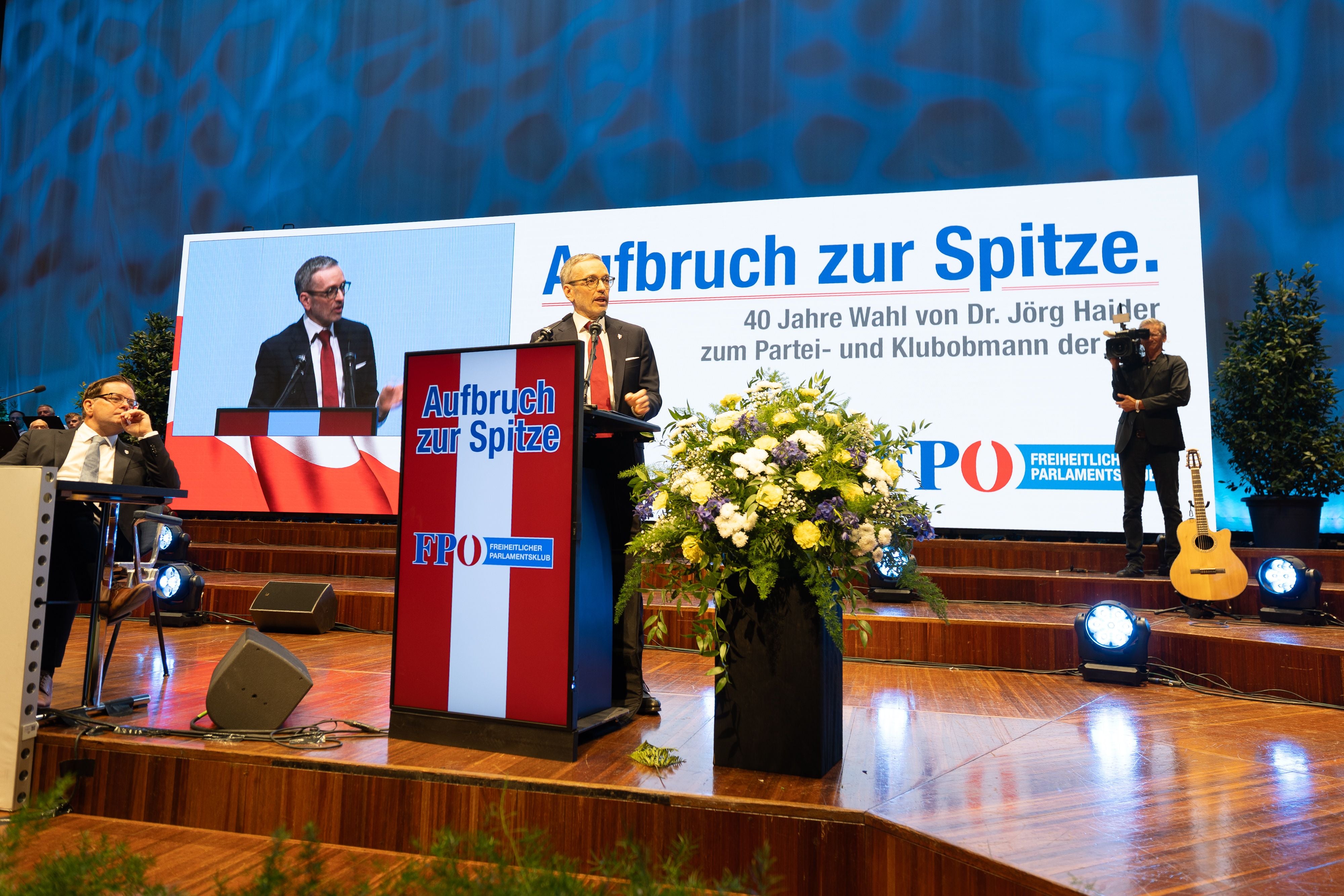 Redner am Podium beim FPÖ-Festakt in Innsbruck, im Hintergrund eine große Jubiläumsleinwand.