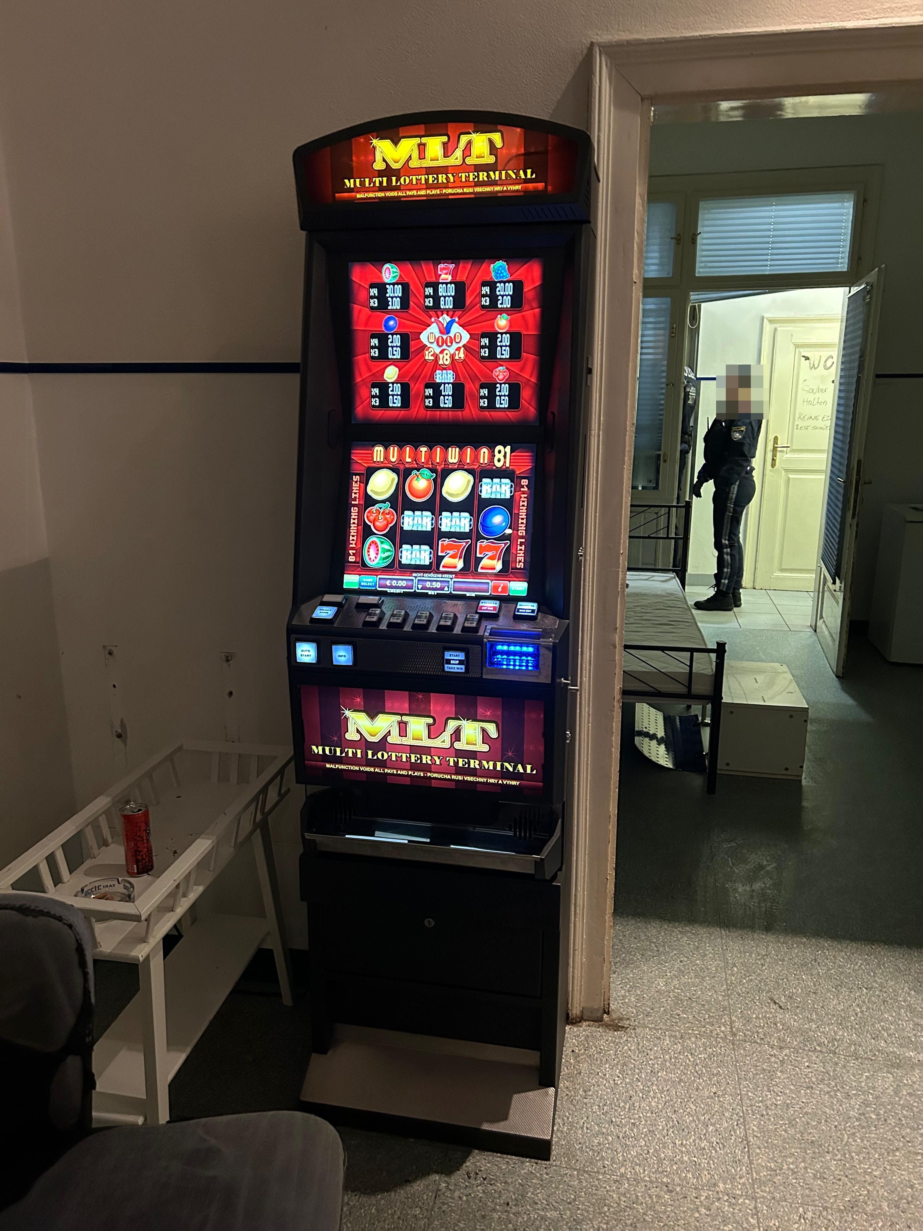 Spielautomat in einem spärlich eingerichteten Raum, im Hintergrund steht eine Polizistin.
