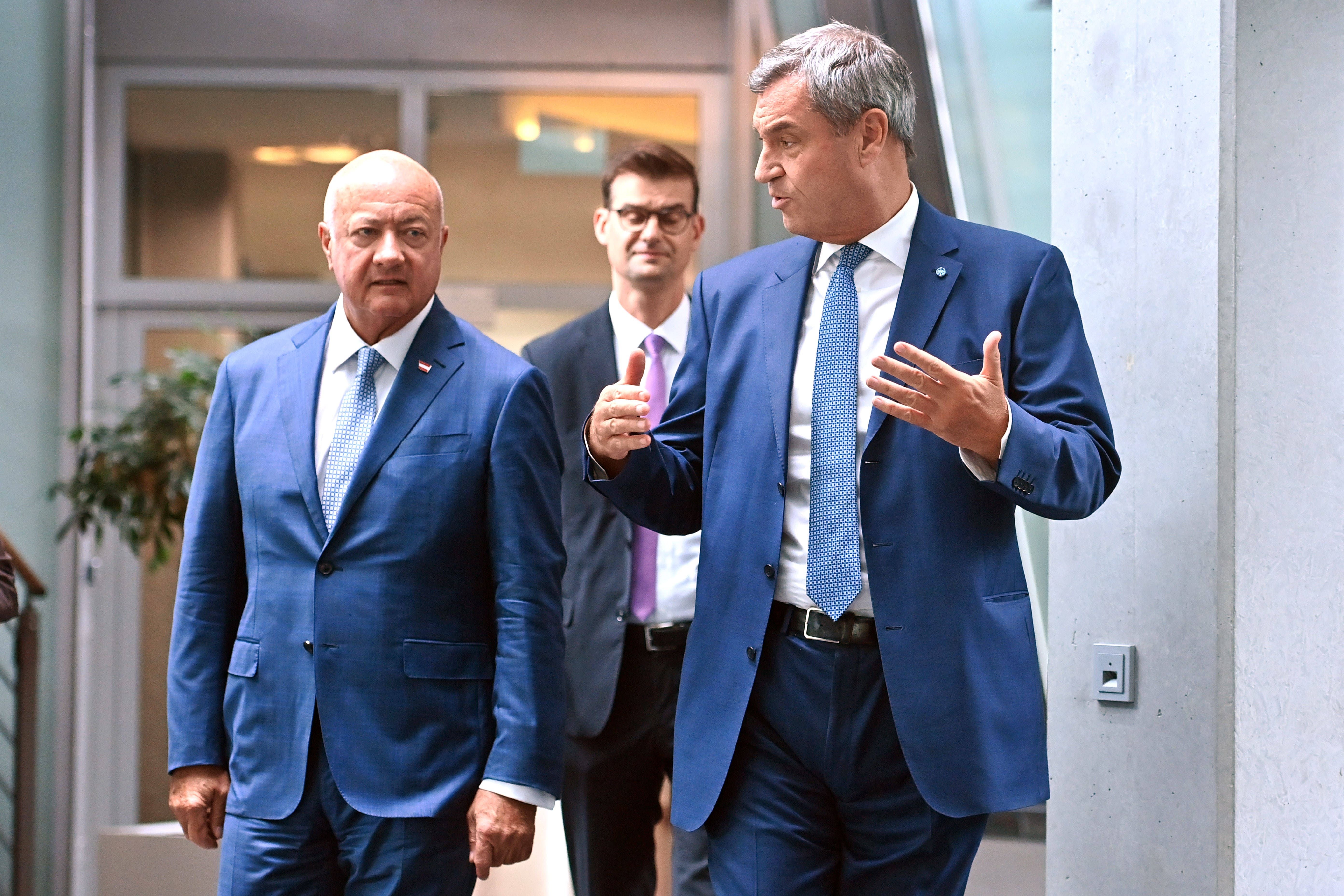 Drei Männer in Anzügen unterhalten sich in einem modernen Gebäude bei einer Pressekonferenz.