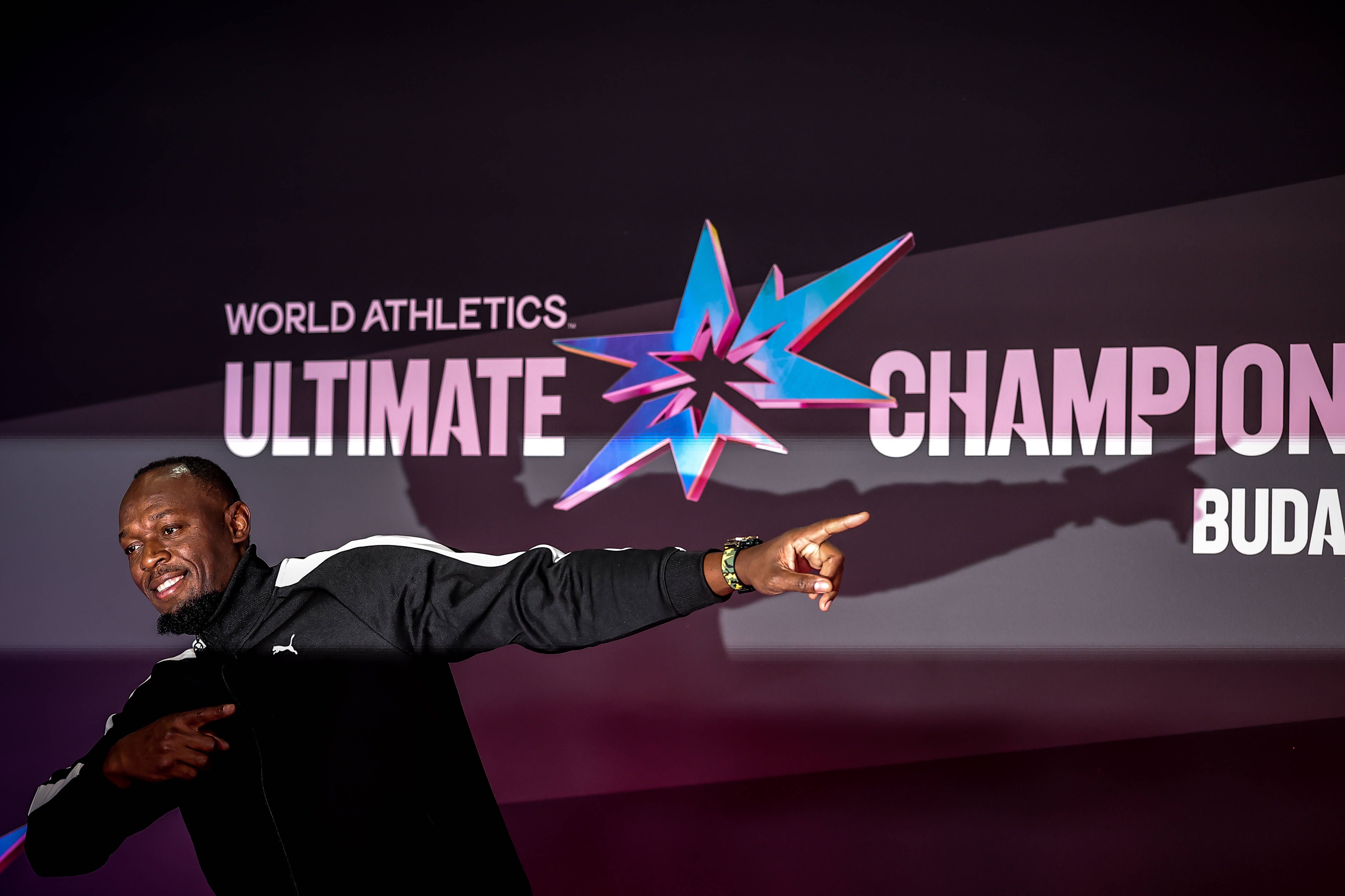 Usain Bolt lächelt und zeigt mit dem Zeigefinger vor der Sponsorenwand der World Athletics Ultimate Championship in Budapest.
