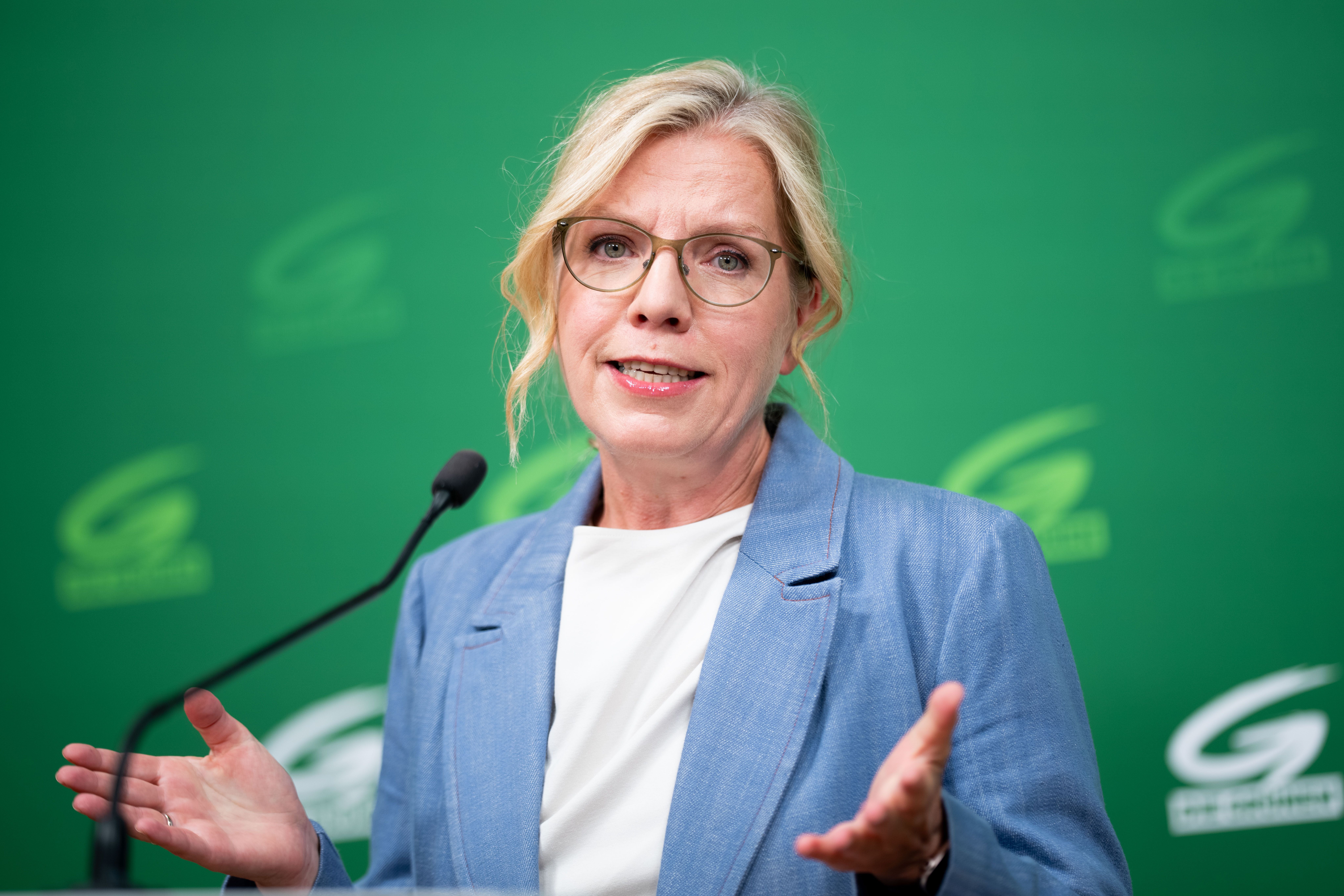 Eine Frau mit Brille spricht bei einer Pressekonferenz vor grünem Hintergrund und Mikrofon.