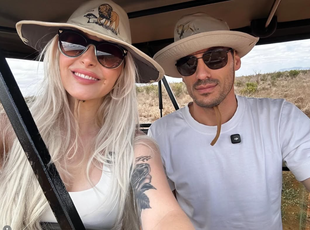 Zwei Personen mit Safarihüten und Sonnenbrillen sitzen in einem offenen Fahrzeug in der Natur.