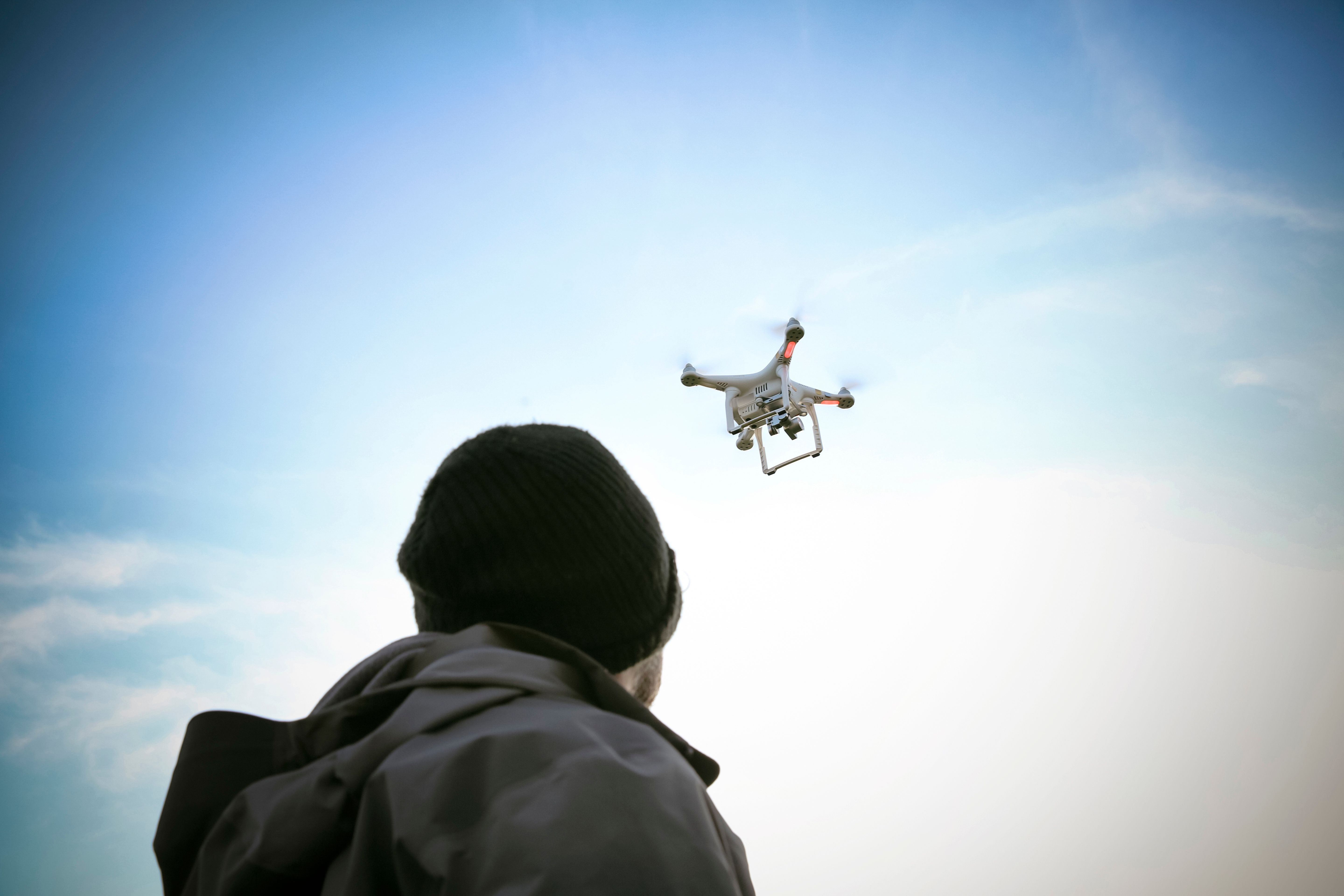 Person mit Mütze beobachtet eine fliegende Drohne am Himmel bei Tageslicht.